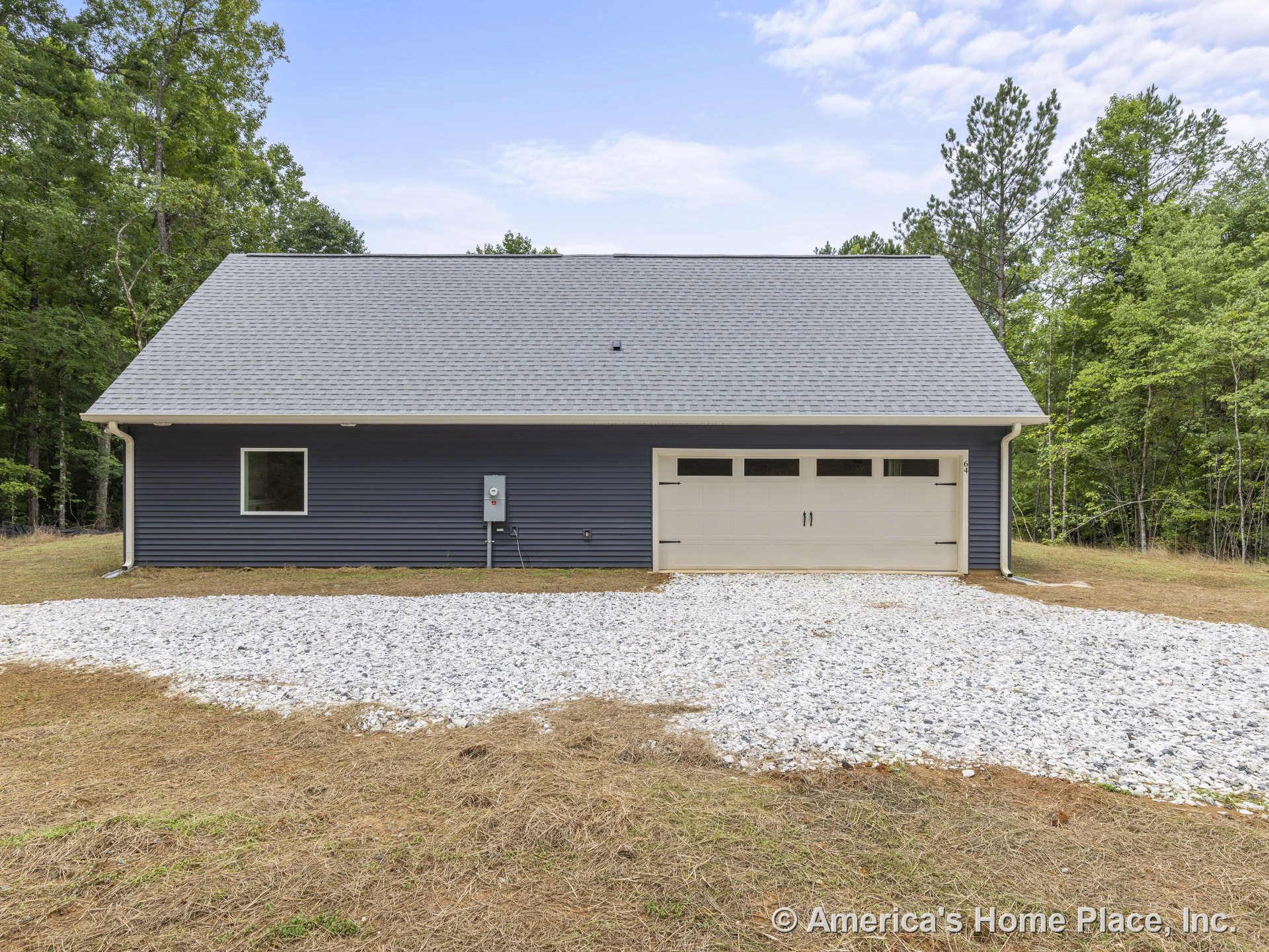Two-car garage with light overhead door, dark horizontal siding, single window, gray shingle roof, gravel driveway, white trim, and exterior utility box in rural setting.