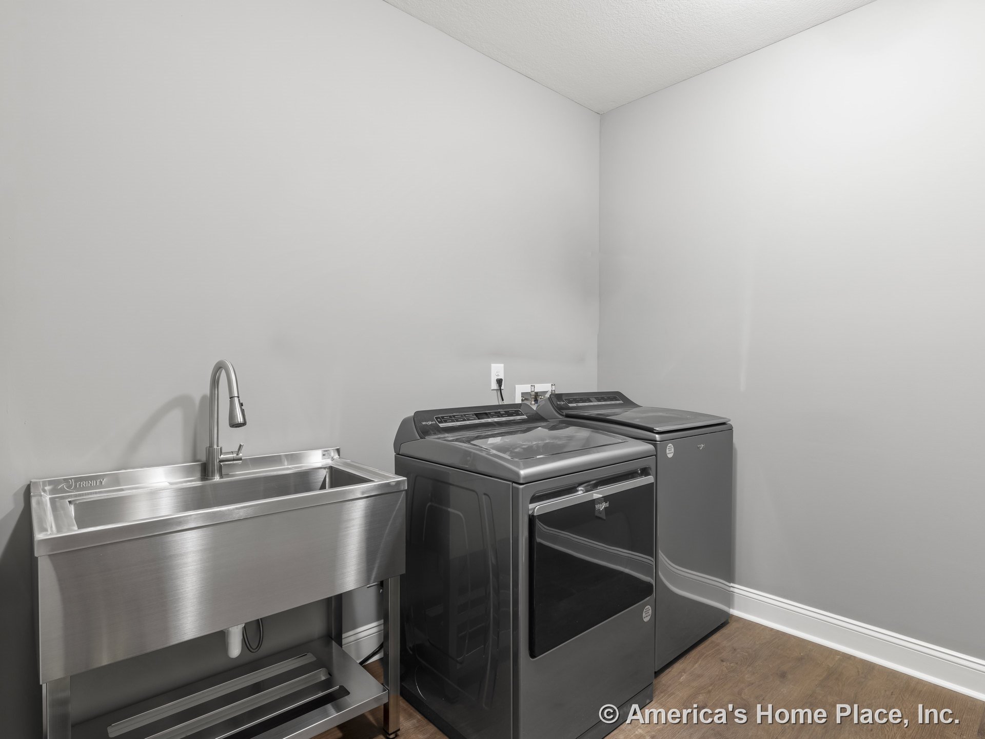 Stainless steel utility sink beside modern front-load washer and dryer, gray painted walls, wood-look flooring, and white baseboard trim.