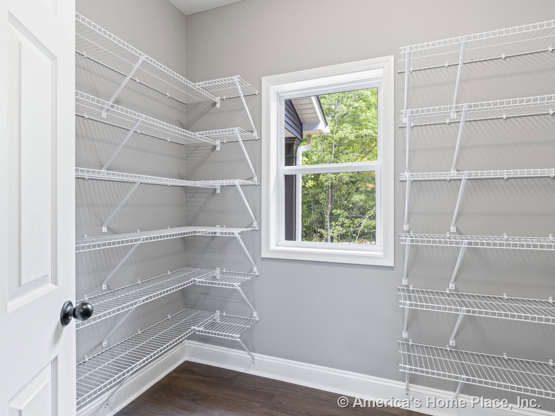 Walk-in pantry with multiple white wire shelving units, gray painted walls, wood-look flooring, window with white trim, and white door featuring a black knob.