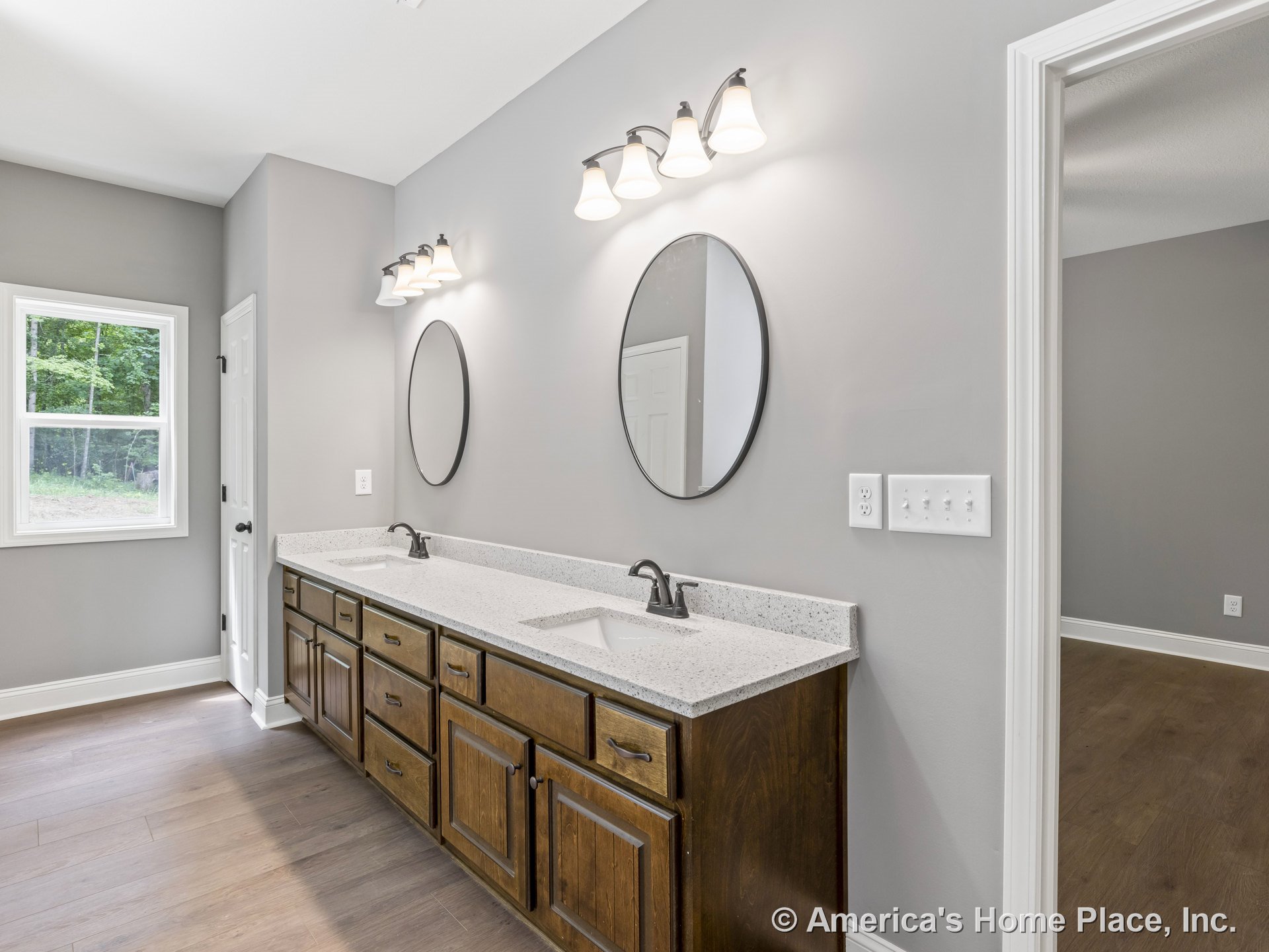 Double vanity with granite countertops and wood cabinetry, two oval mirrors beneath wall-mounted light fixtures, gray painted walls, window with white trim, door, and tile