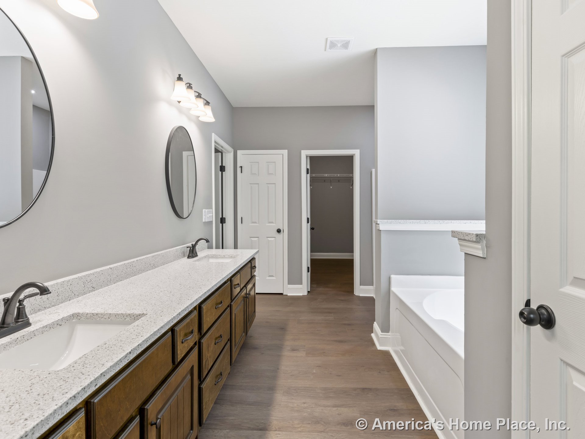 Double vanity with quartz countertops and wood cabinetry, built-in bathtub with tile surround, round wall mirrors above sinks, wall-mounted vanity lighting, walk-in closet with