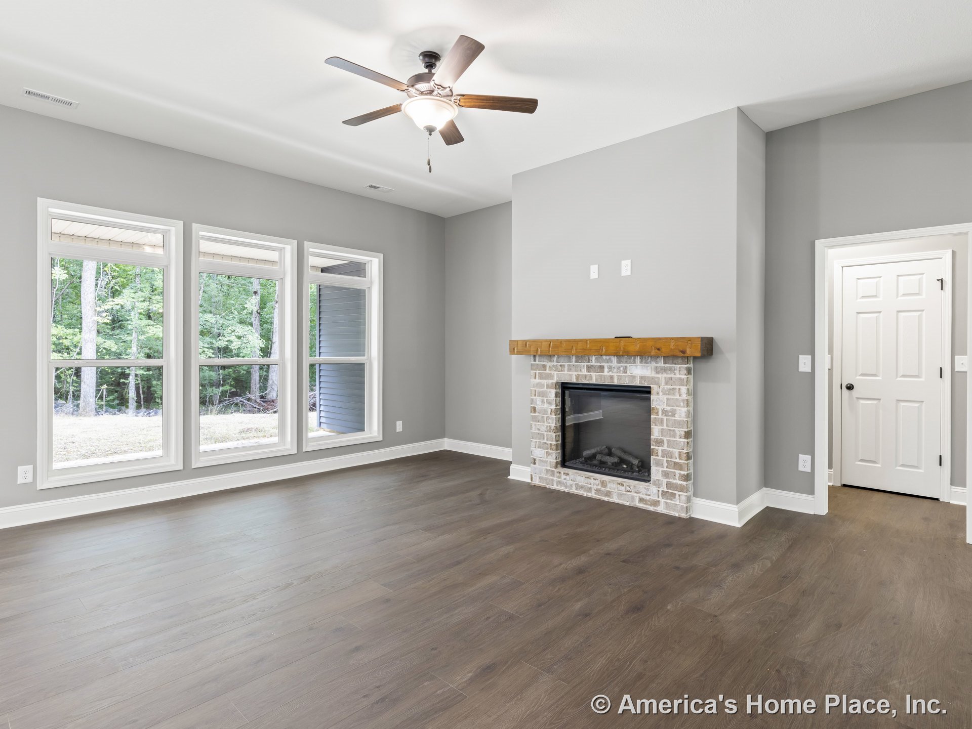 Brick fireplace with wood mantel centered on gray painted living room wall, wide plank wood flooring, large triple windows with white trim and baseboards, ceiling fan with light