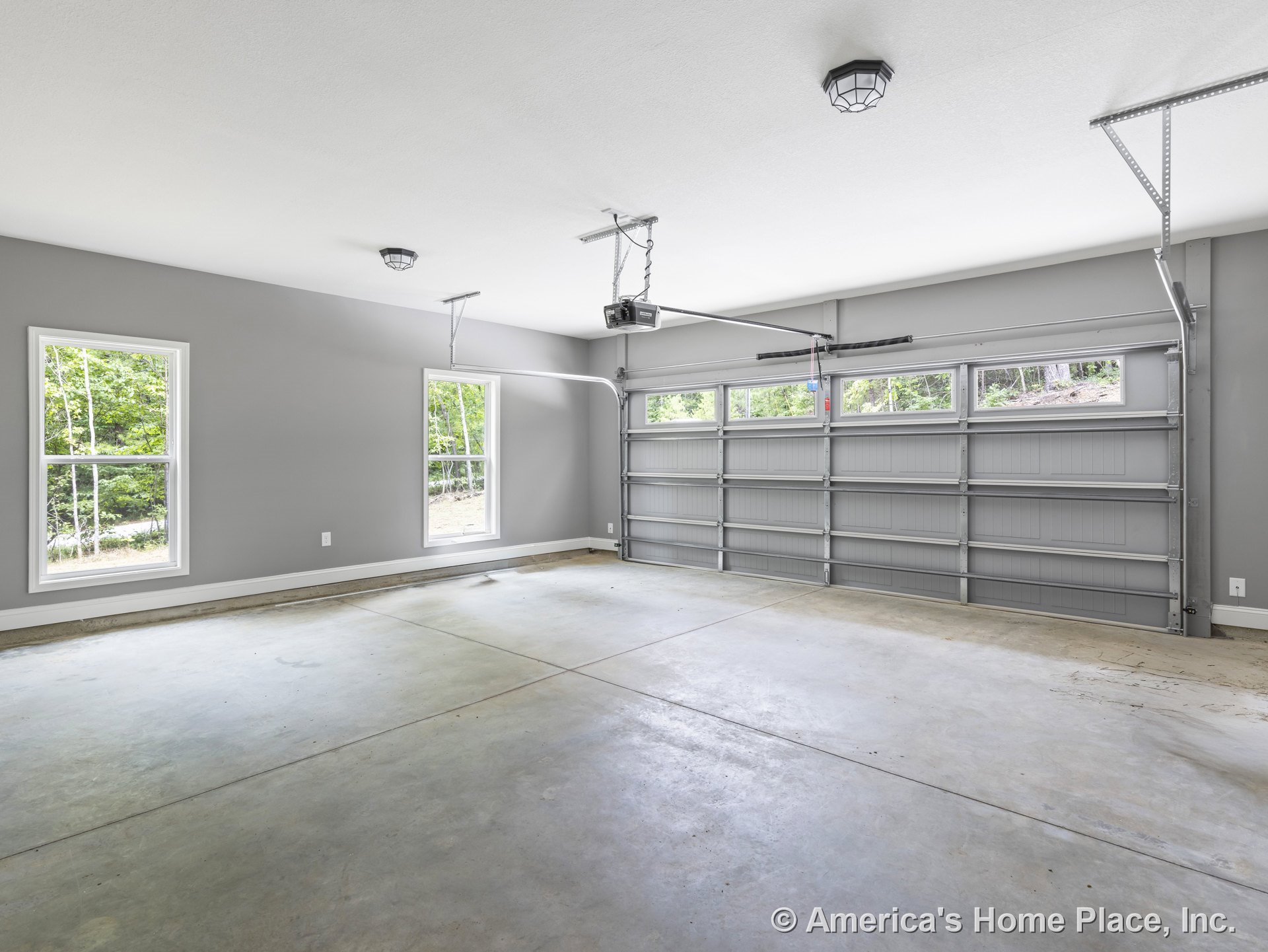 Finished garage with gray painted walls, concrete slab flooring, sectional garage door featuring upper windows, two double-hung windows, ceiling-mounted light fixtures, and an
