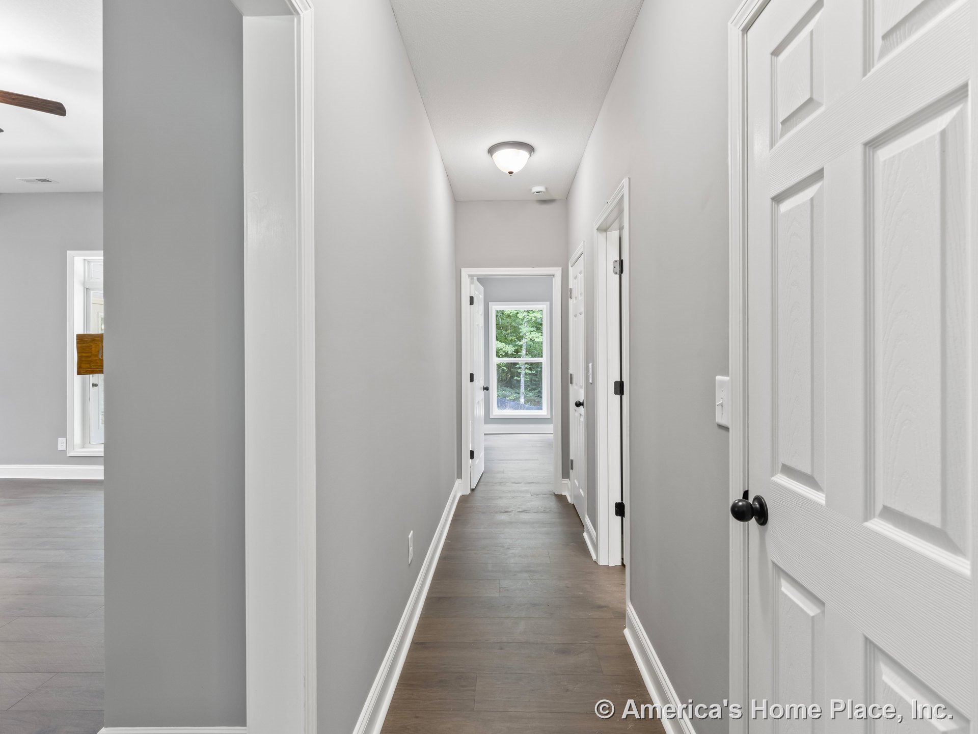 Hallway with light gray painted walls, white baseboards and trim, white paneled doors, wood-look plank flooring, flush mount ceiling light, and large window at the end providing