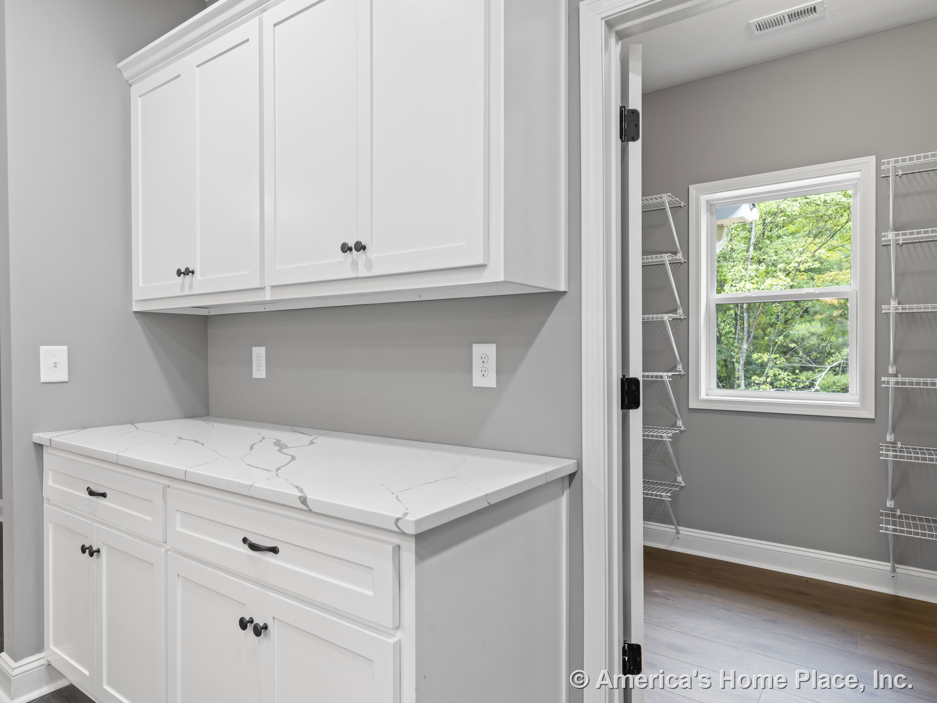 White upper and lower pantry cabinets with marble-look countertops, wire shelving, gray painted walls, window with trim, and wood flooring.