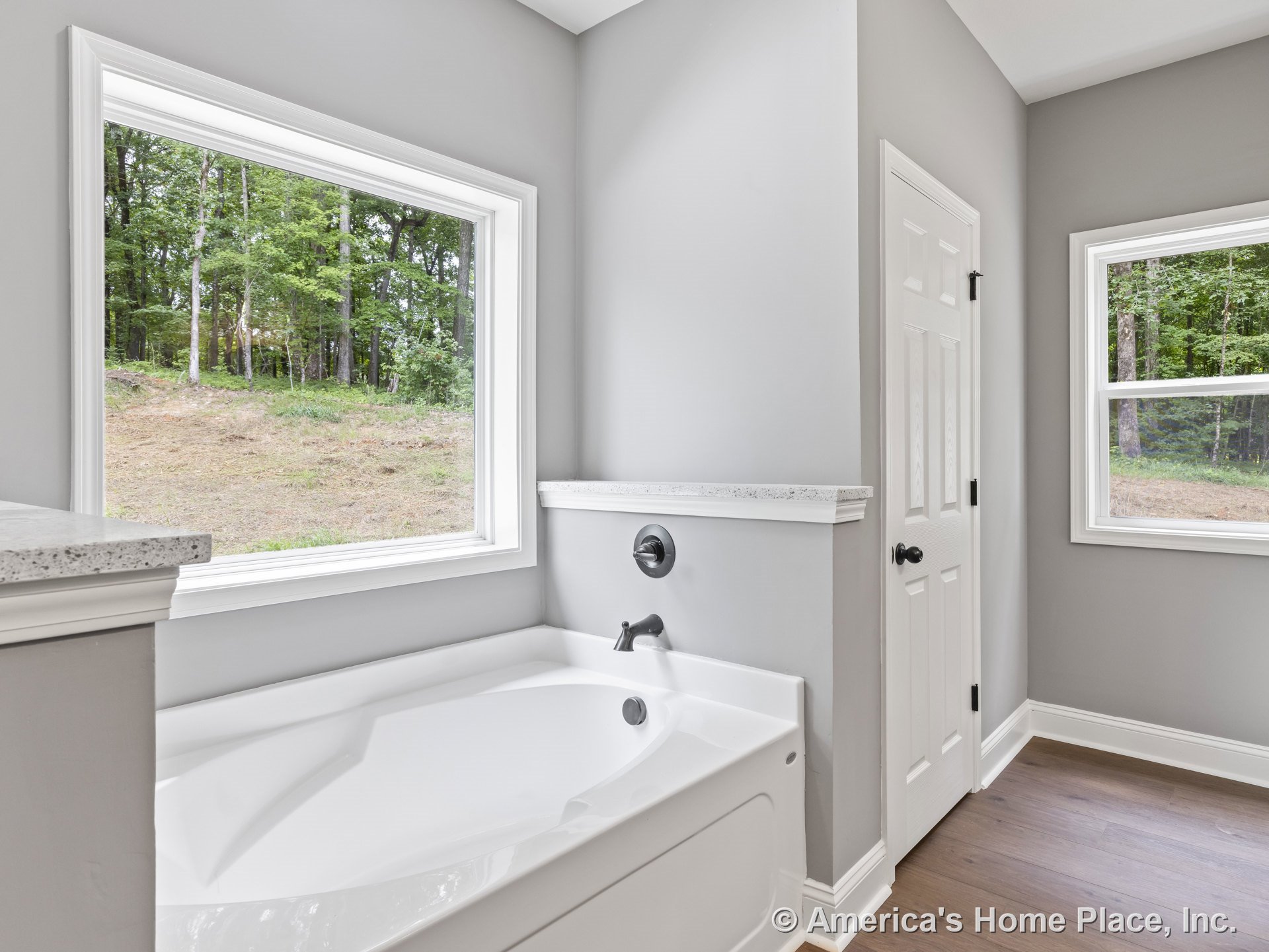 Built-in soaking tub with chrome faucet positioned beneath a large picture window framed in white wood trim, gray painted walls, wood-look flooring, and panel door with black