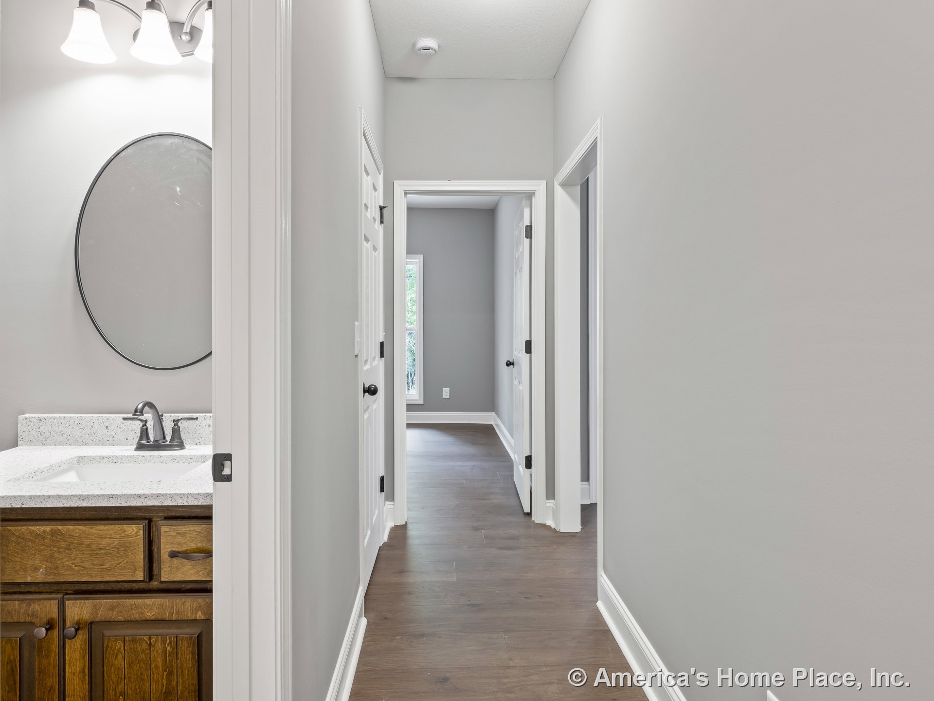 Hallway with wood flooring, white trim and interior doors, built-in overhead lighting, neutral wall paint; bathroom on left features quartz countertop vanity, round wall-mounted