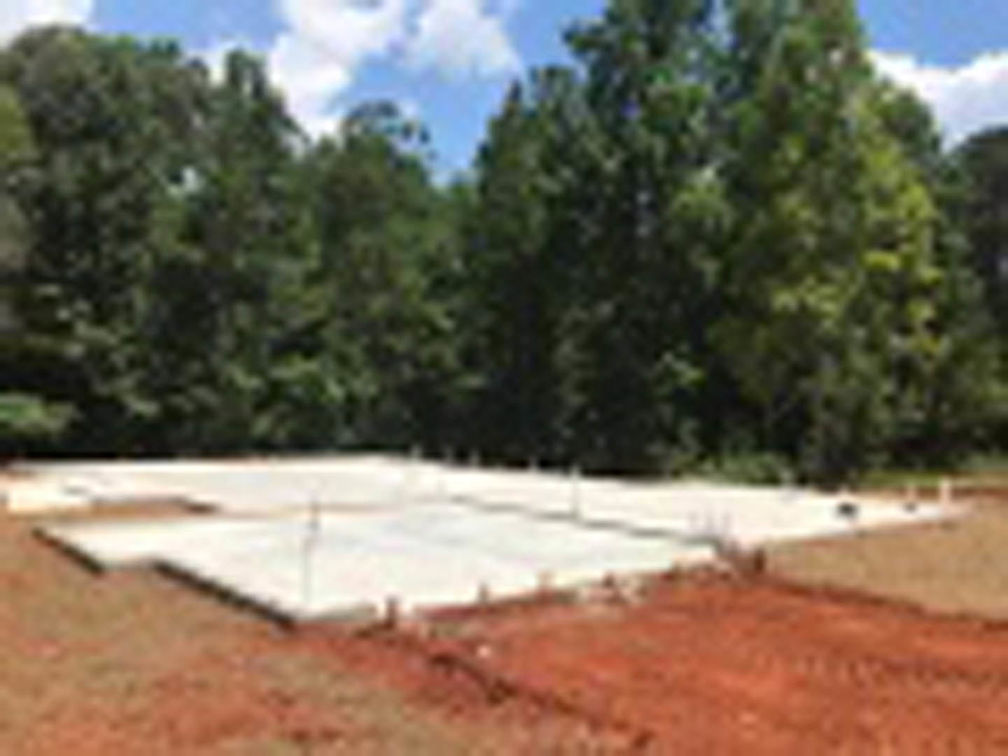 Concrete foundation slab set in grassy field, bordered by mature trees under clear blue sky