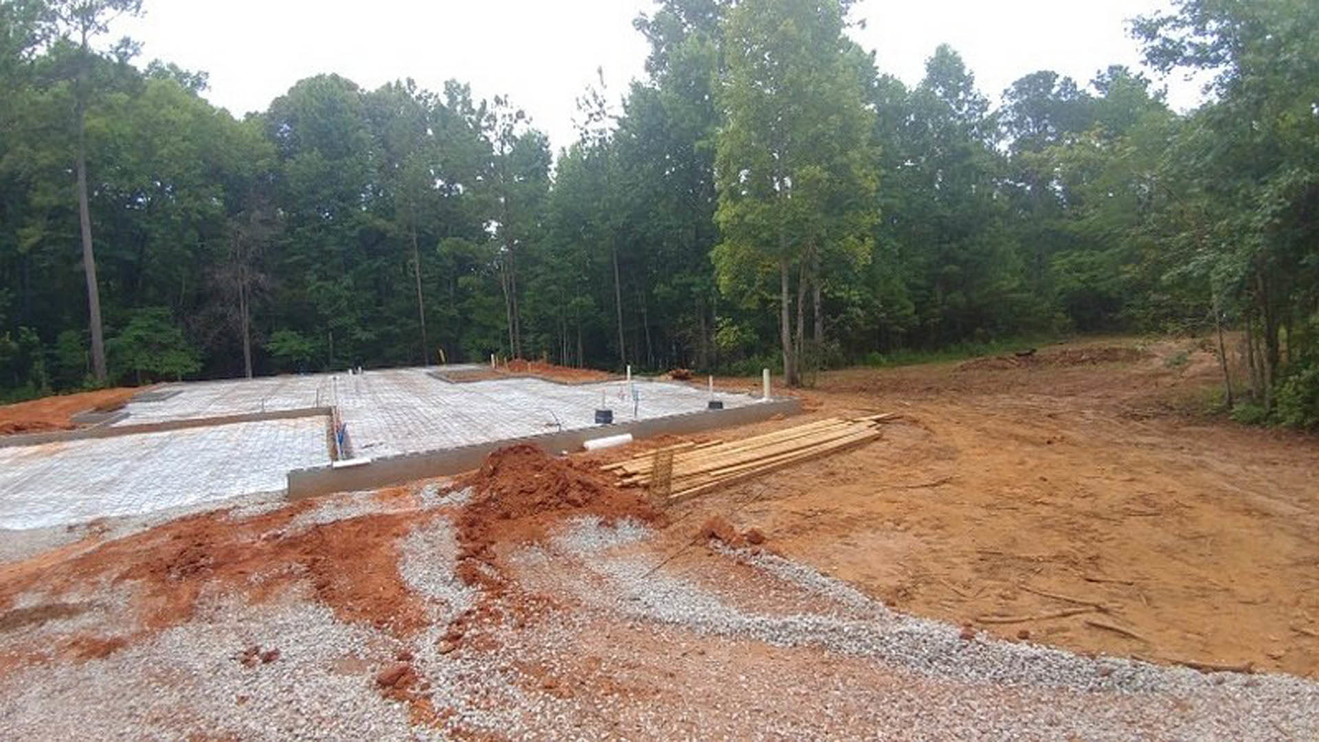 Wood framing and gravel piles on a residential construction site bordered by leafy trees and exposed soil