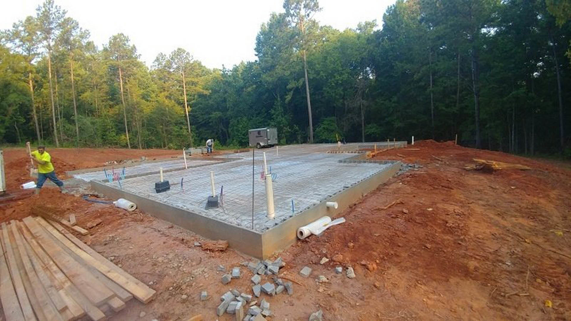 Concrete foundation with exposed pipes and poles surrounded by dirt, piles of wood and broken bricks, construction trailer with logo, man in yellow shirt, trees in background
