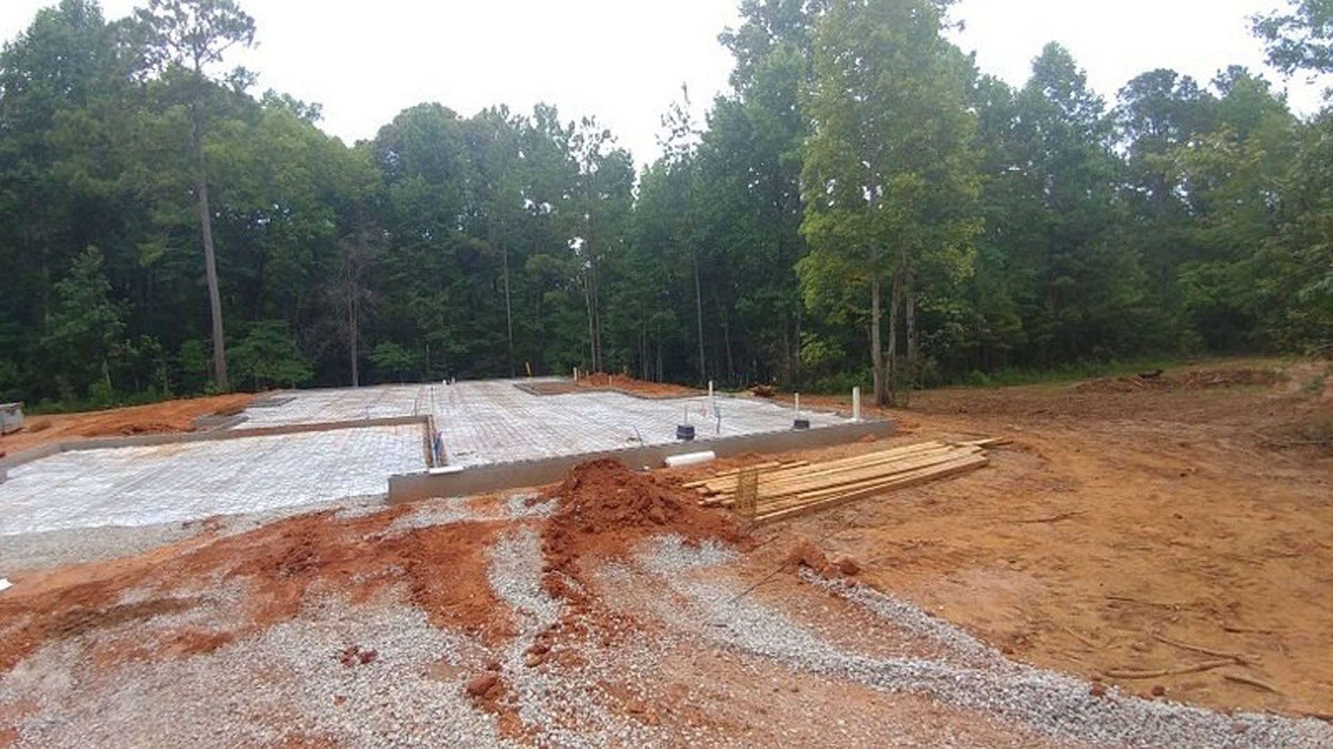 Framed foundation and piles of wood on a dirt construction site, surrounded by tall trees and cloudy sky in the background