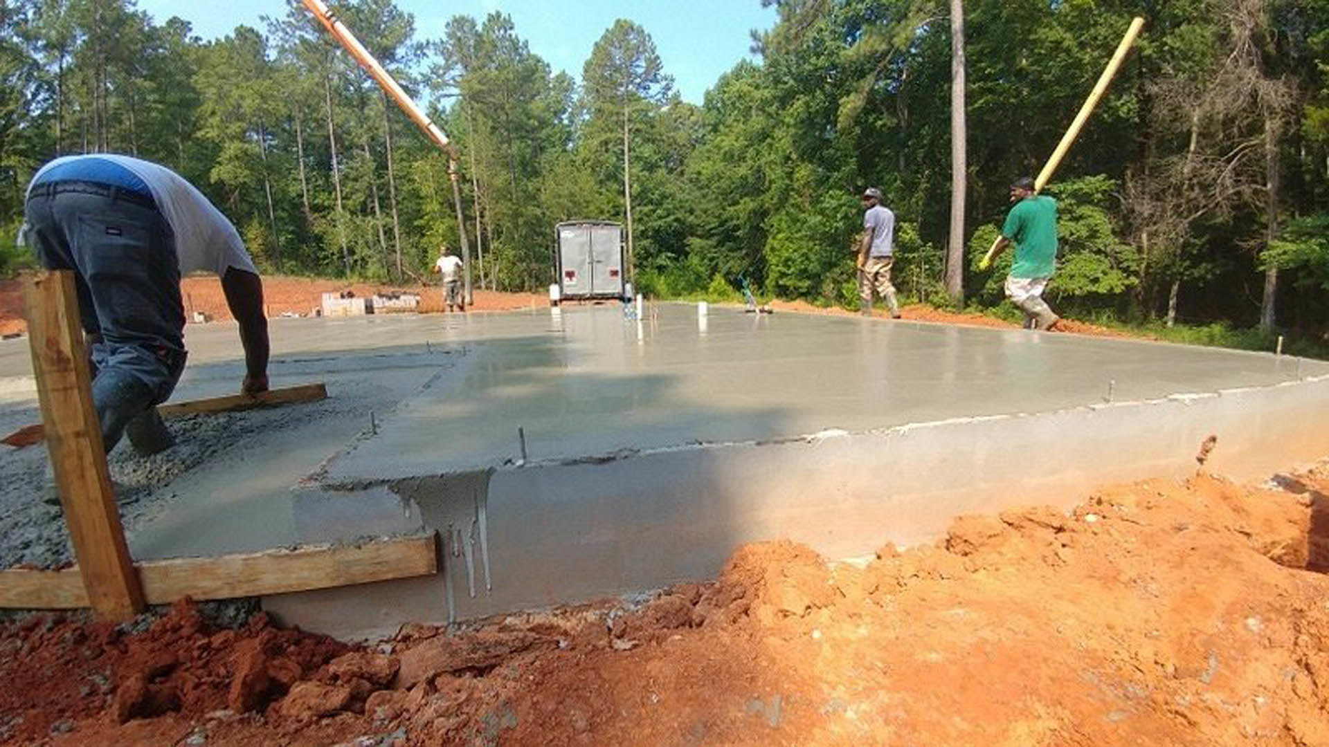 Several men standing on a newly poured concrete foundation surrounded by trees, with construction materials and a white trailer visible in the background.