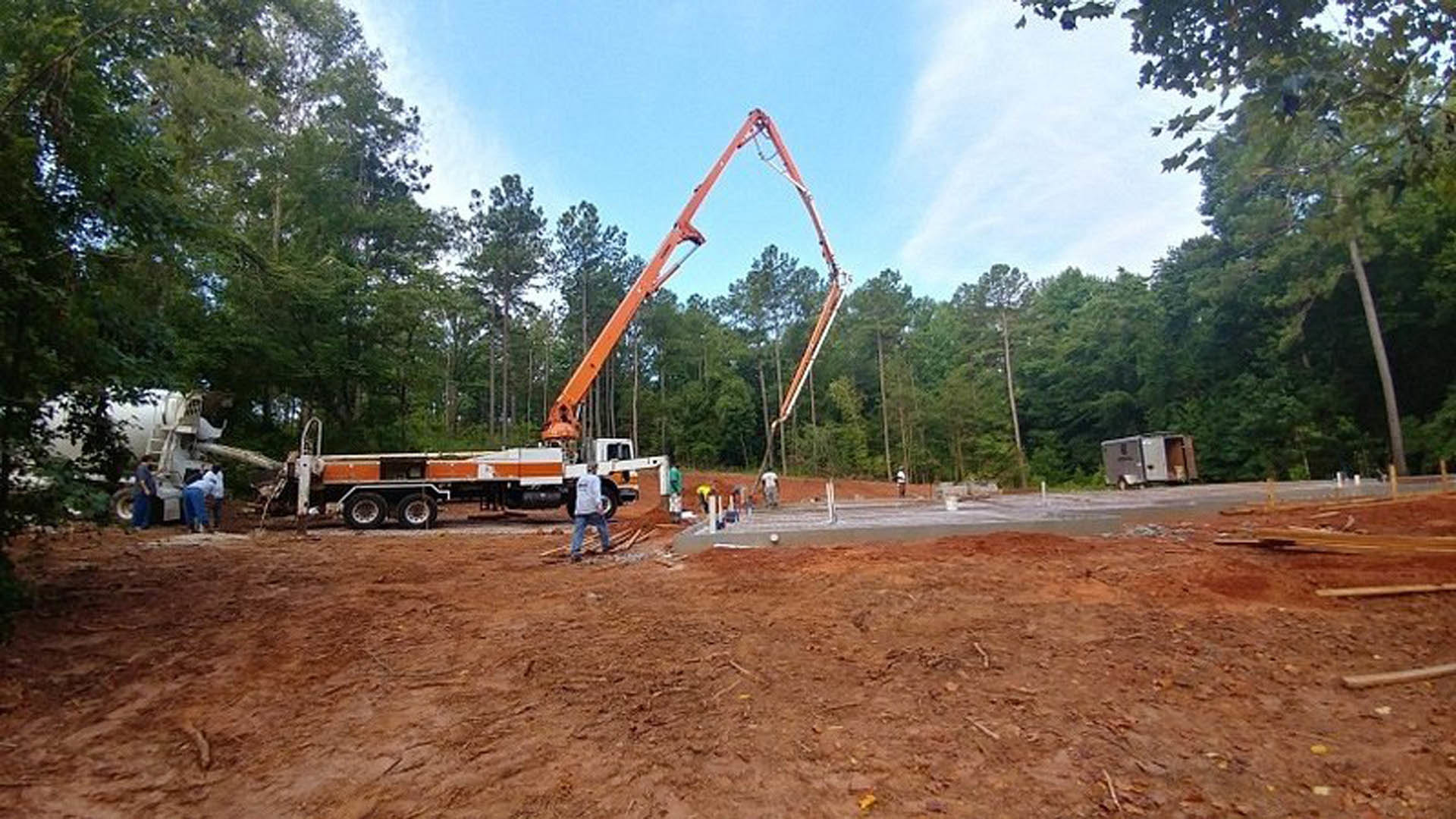 Crane hoisting concrete slab above dirt lot, white trailer with black text parked nearby, trees and construction equipment visible under clear sky