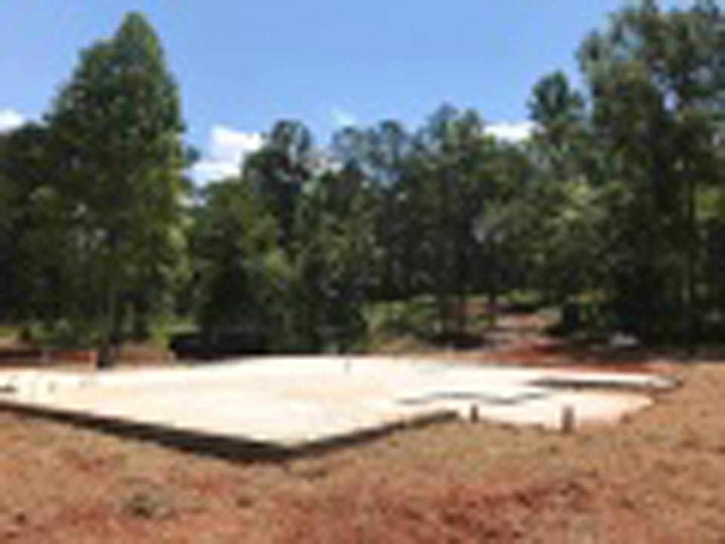 Concrete foundation slab set in grassy field, bordered by tall trees under clear blue sky