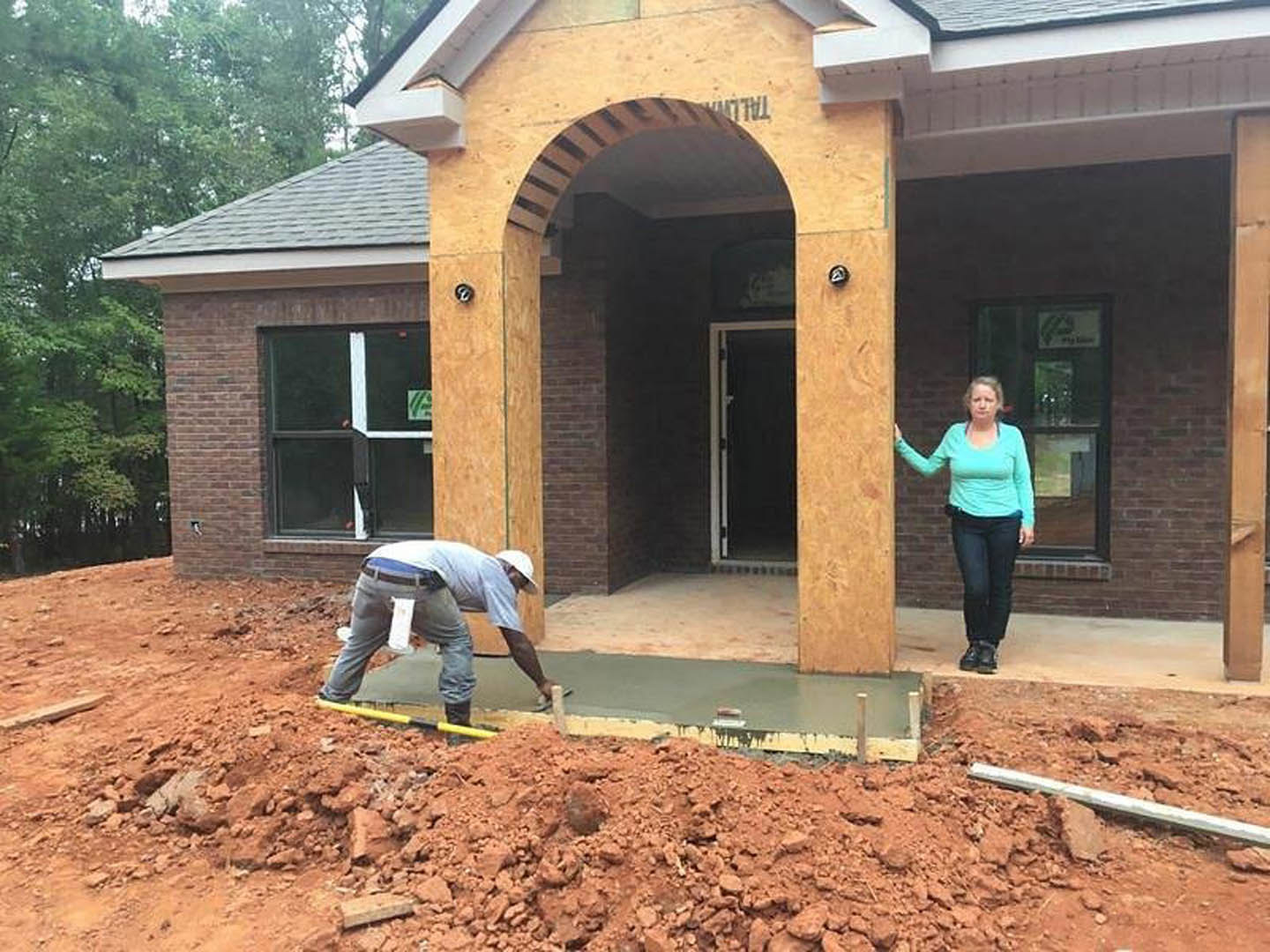 Two people standing on soil in front of a modern house with black door, white trim, large window, and tree nearby