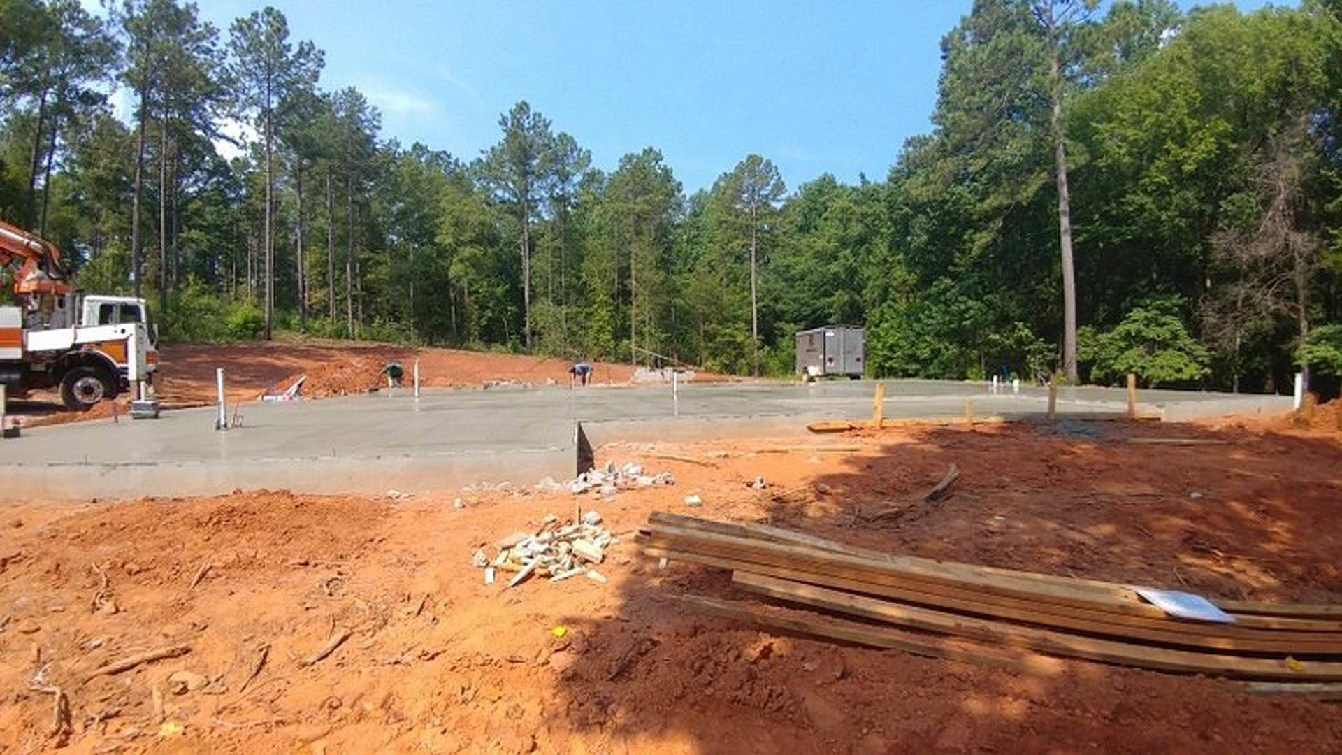 Framed custom home under construction with exposed wood beams, construction truck parked on dirt lot, piles of lumber and materials scattered, mature trees lining background