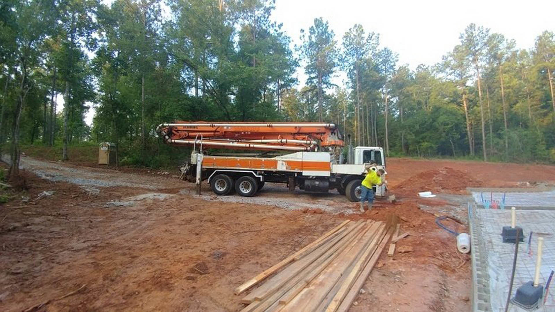 Construction site with a white truck parked on dirt, large metal object loaded in the bed, scattered wood piles nearby, trees in the background, and a person in a yellow shirt