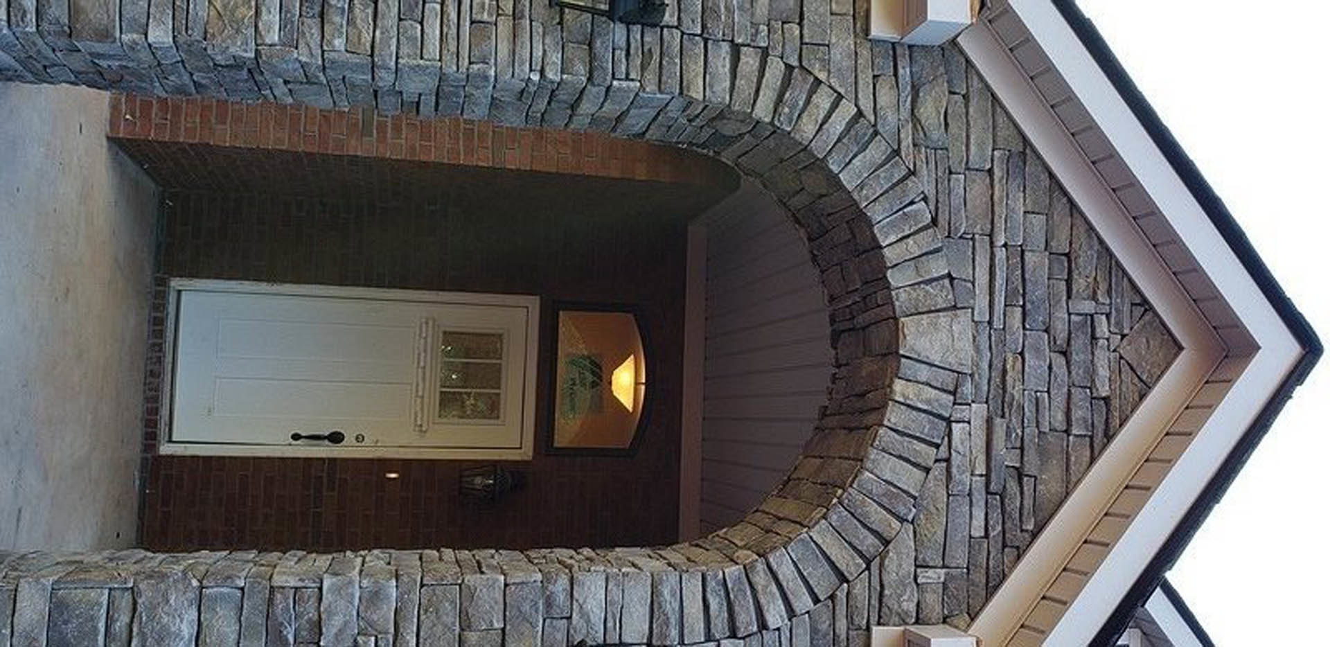 Stone archway framing a white door with black handle, exterior wall of textured stone, small light fixture above doorway, blurred green plant in foreground