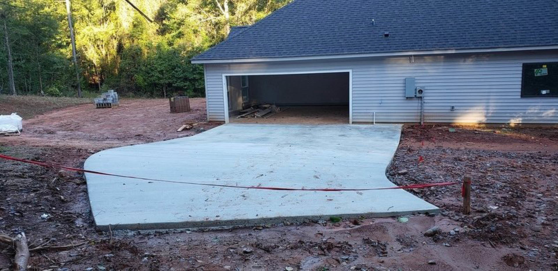 Garage with open door, blue siding and white trim, concrete driveway showing visible crack, surrounded by trees and plants in backyard.