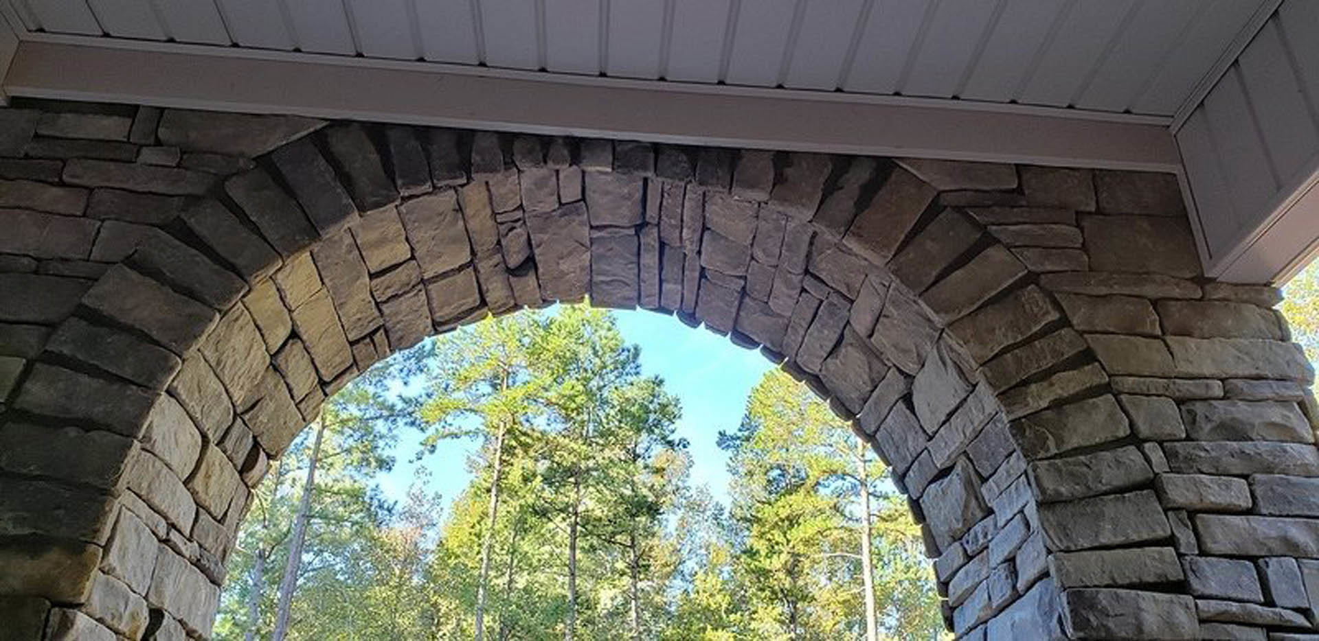 Stone archway framed by tall green trees, brick wall with white shelf, textured stone and white wall details, grey square accent on brick exterior