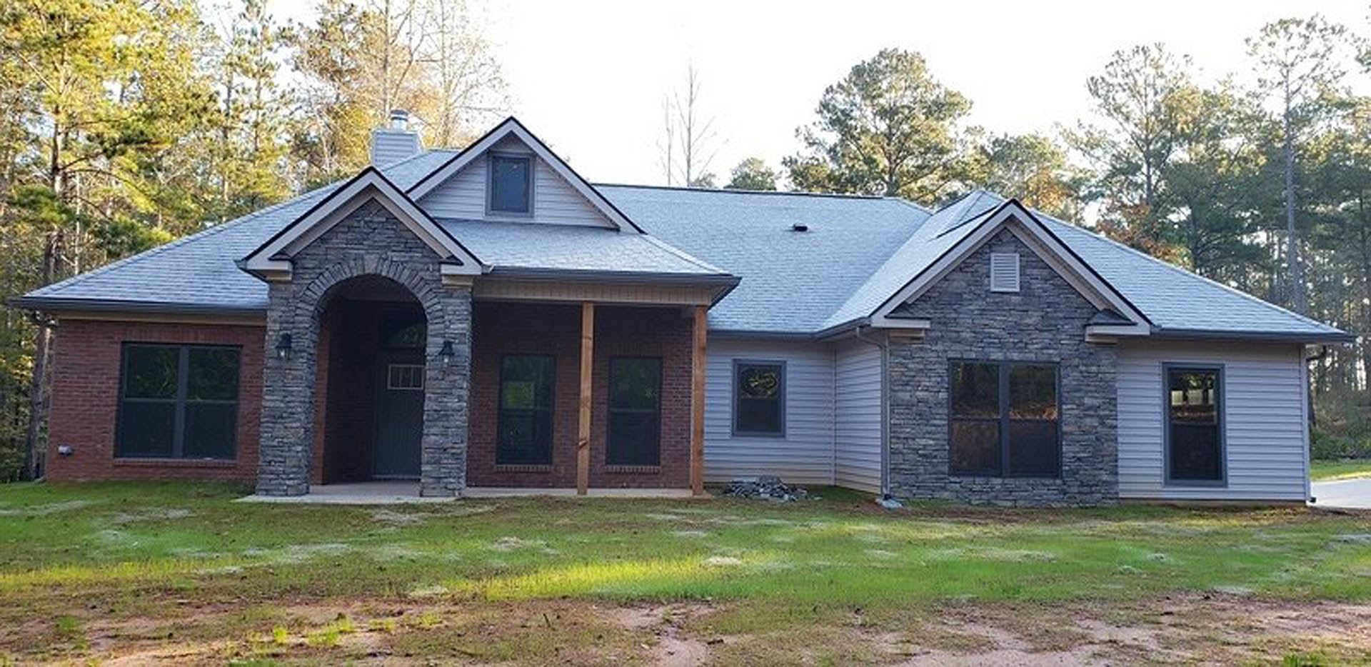 Stone and brick exterior walls, blue-framed window, dark-framed window, porch, vent on brick wall, surrounded by grass and trees under a clear sky