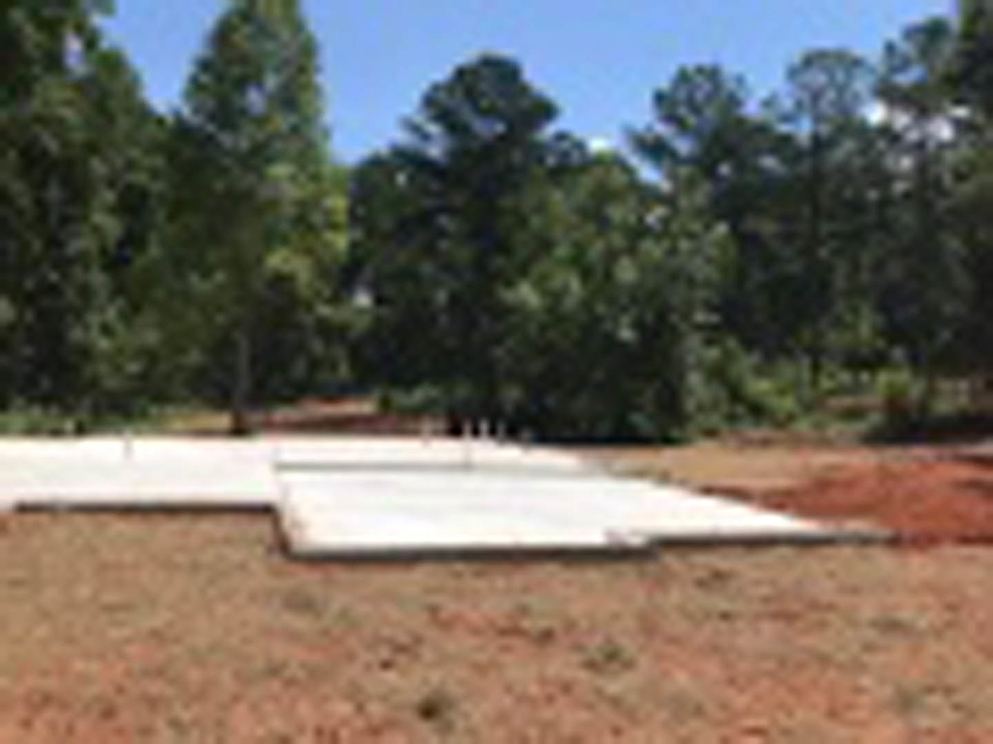 Concrete slab foundation set in a grassy field with mature trees in the background and a dirt road nearby