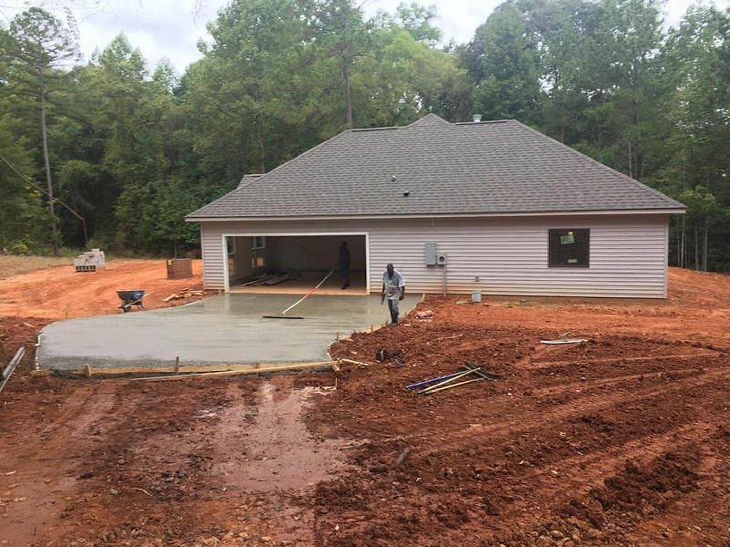 Modern home with attached garage, concrete driveway, brick exterior walls, black garage door, small trees and plants along the edge, man walking toward garage, stack of boxes near