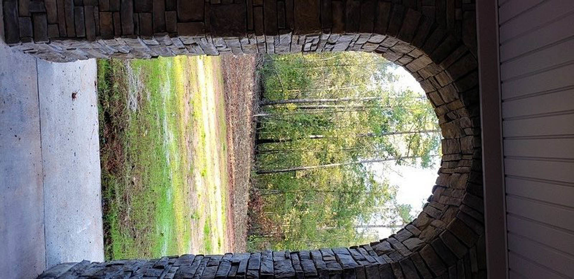 Stone archway framed by mature trees, dirt road bordered by grass, textured concrete and white walls, forested backdrop