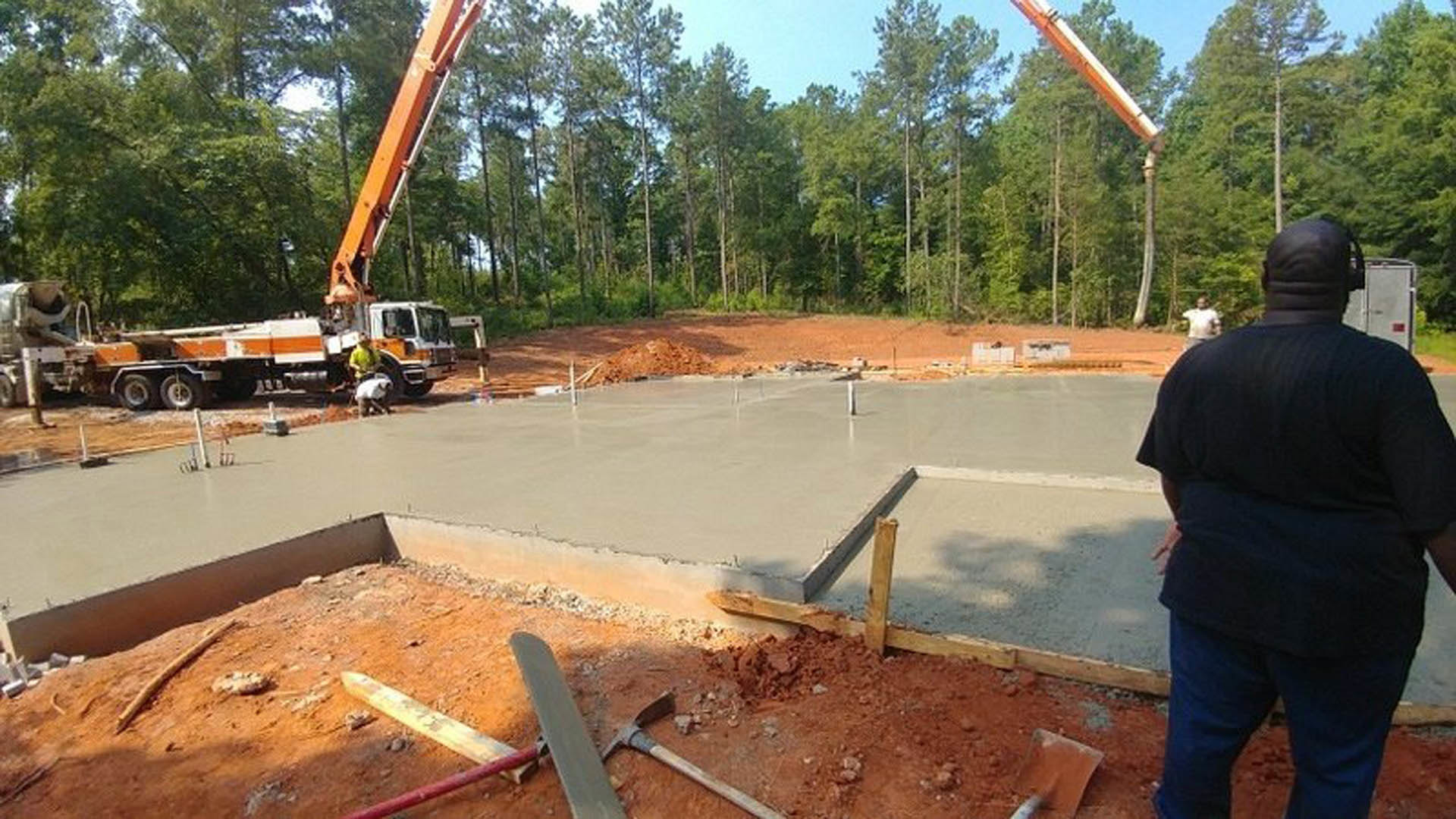 Framed house foundation surrounded by dirt, construction crane, scattered trees, and workers in safety gear