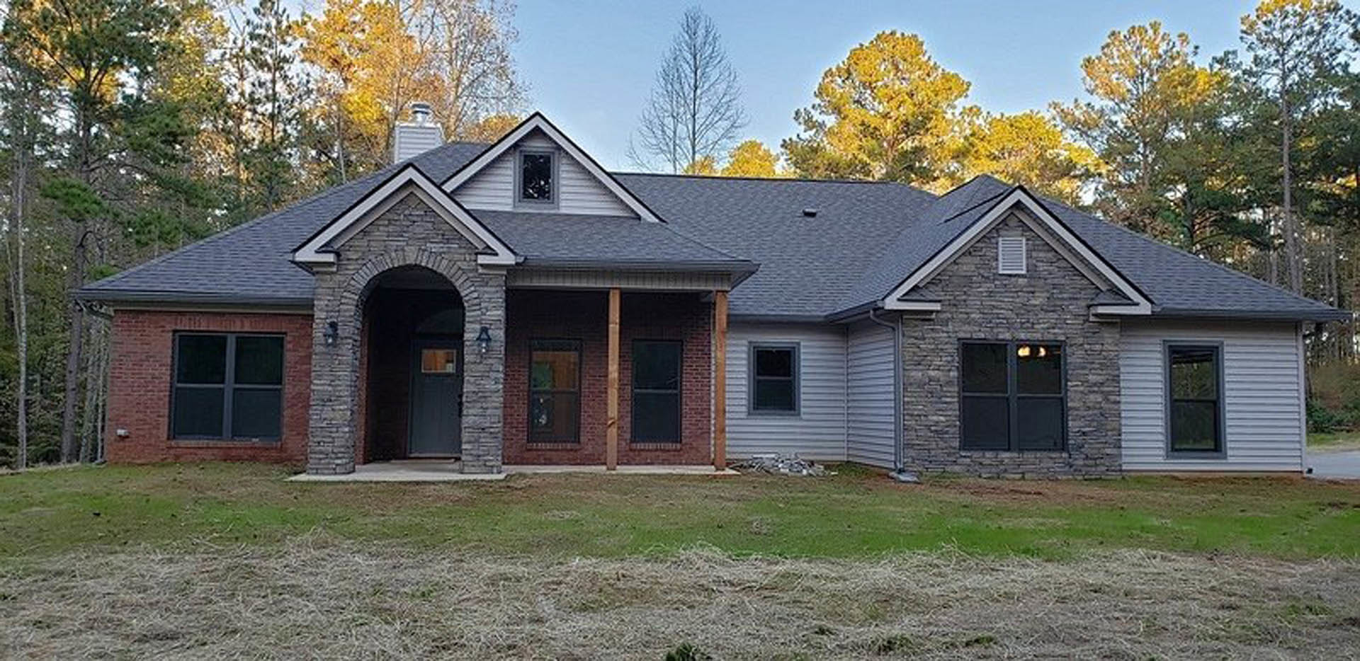 Two-story house with gray siding, black-framed windows, covered front porch, manicured lawn, mature trees in the background, and white vent near the roofline