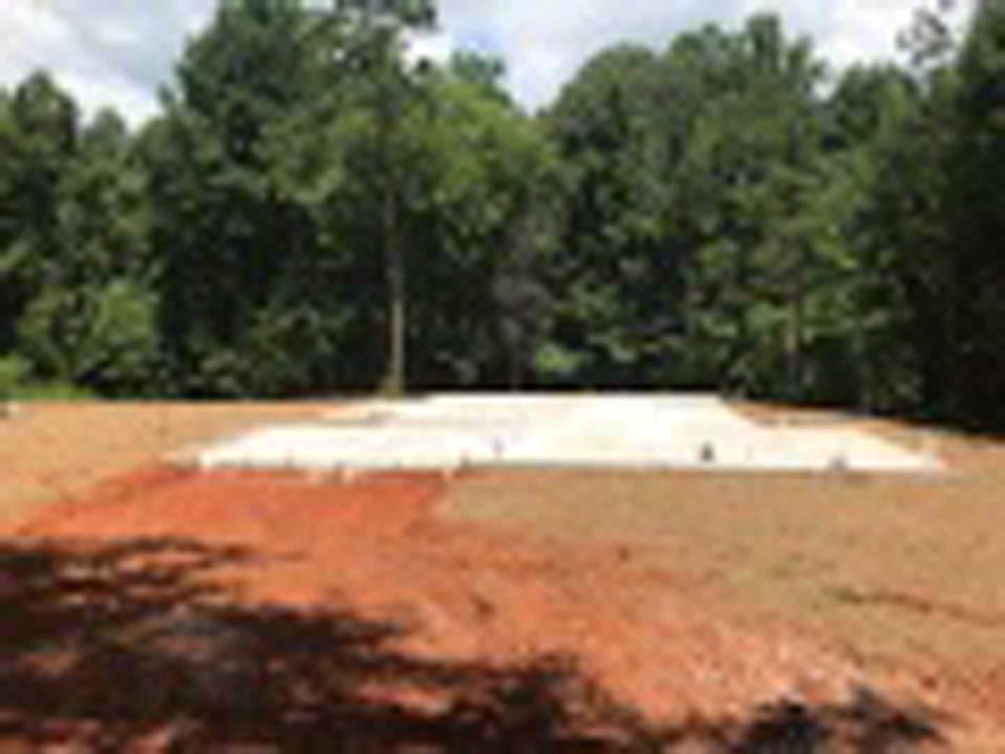 Red dirt field bordered by tall green trees under a pale sky
