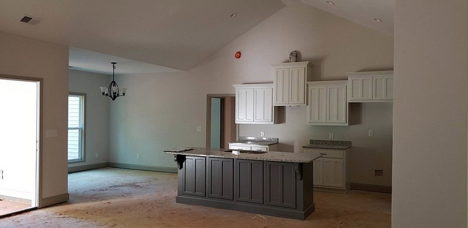 Spacious kitchen featuring a large granite island with built-in sink, white cabinetry, stainless steel appliances, pendant chandelier, hardwood flooring, and tall windows.