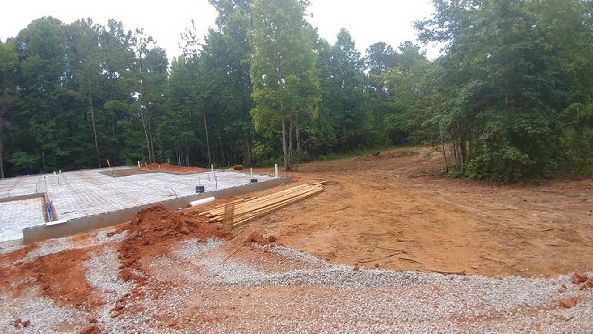 Dirt construction site bordered by tall trees, piles of wood and gravel scattered on ground, concrete slab partially visible under sunlight