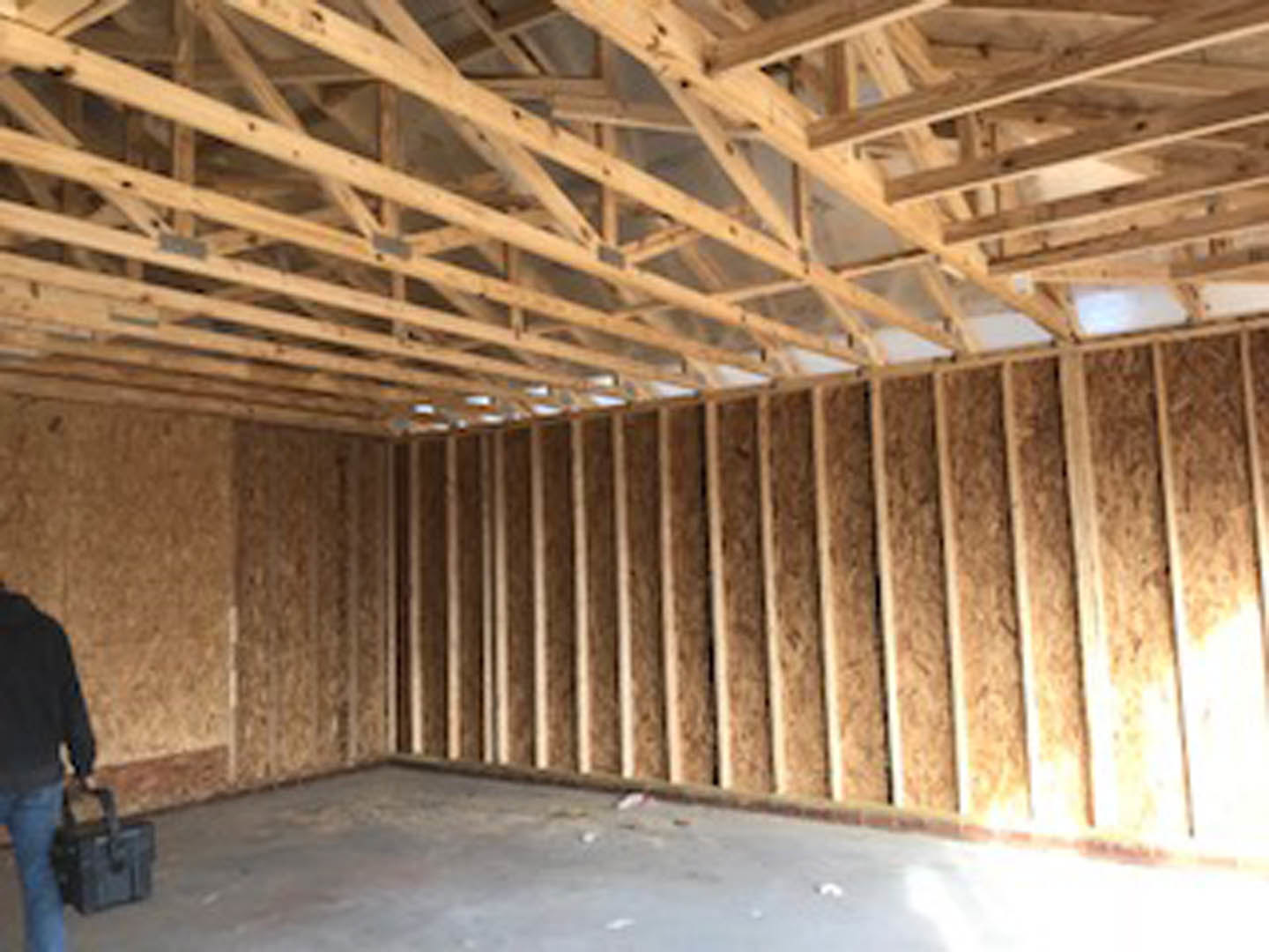 Open room with exposed wooden ceiling beams, wood-framed walls, concrete floor, and a person holding a suitcase partially visible in the foreground.