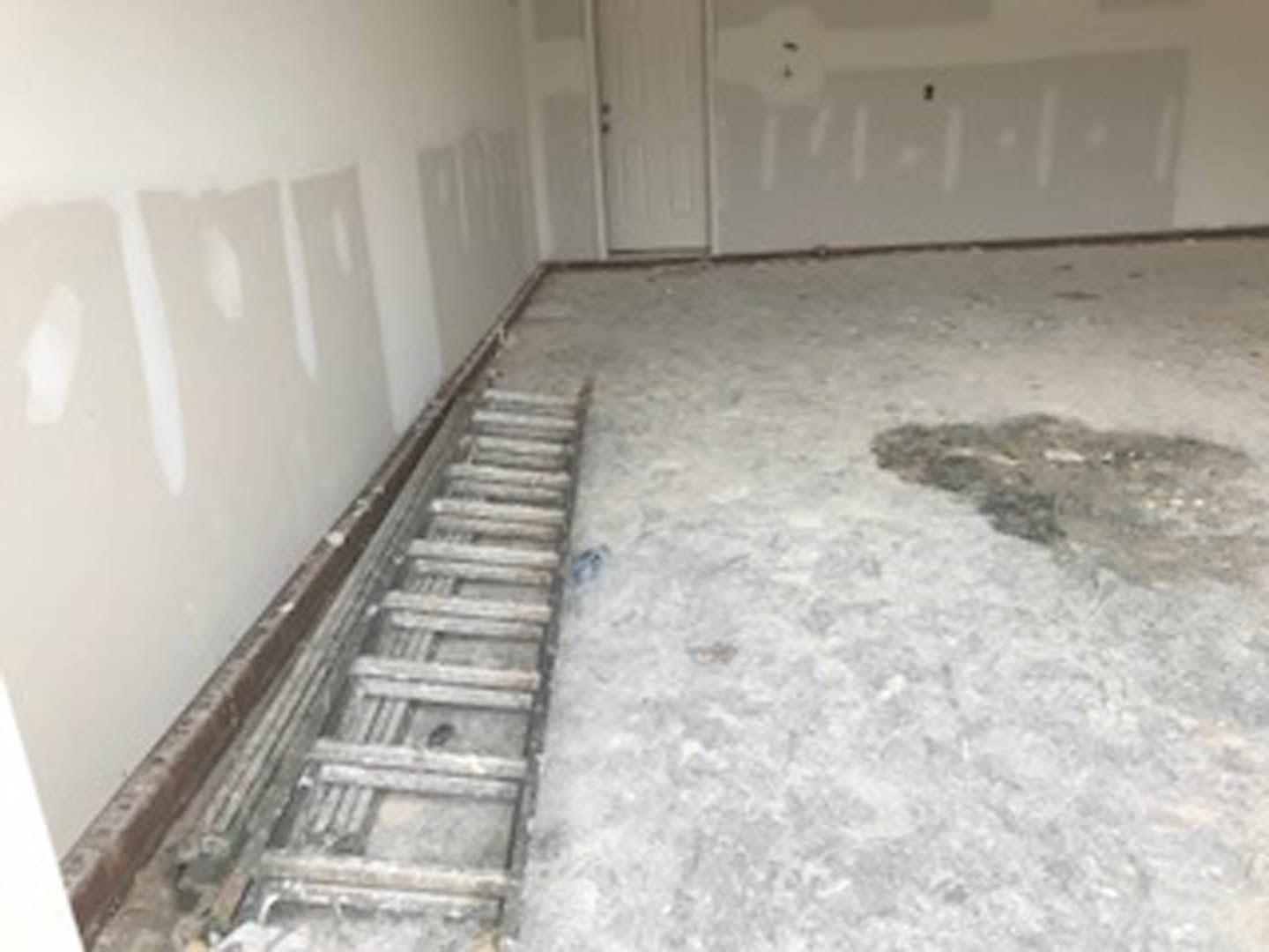 Aluminum ladder standing on unfinished concrete floor with metal grate, white plaster walls, white ceiling with recessed black fixtures, and white door with matching knob