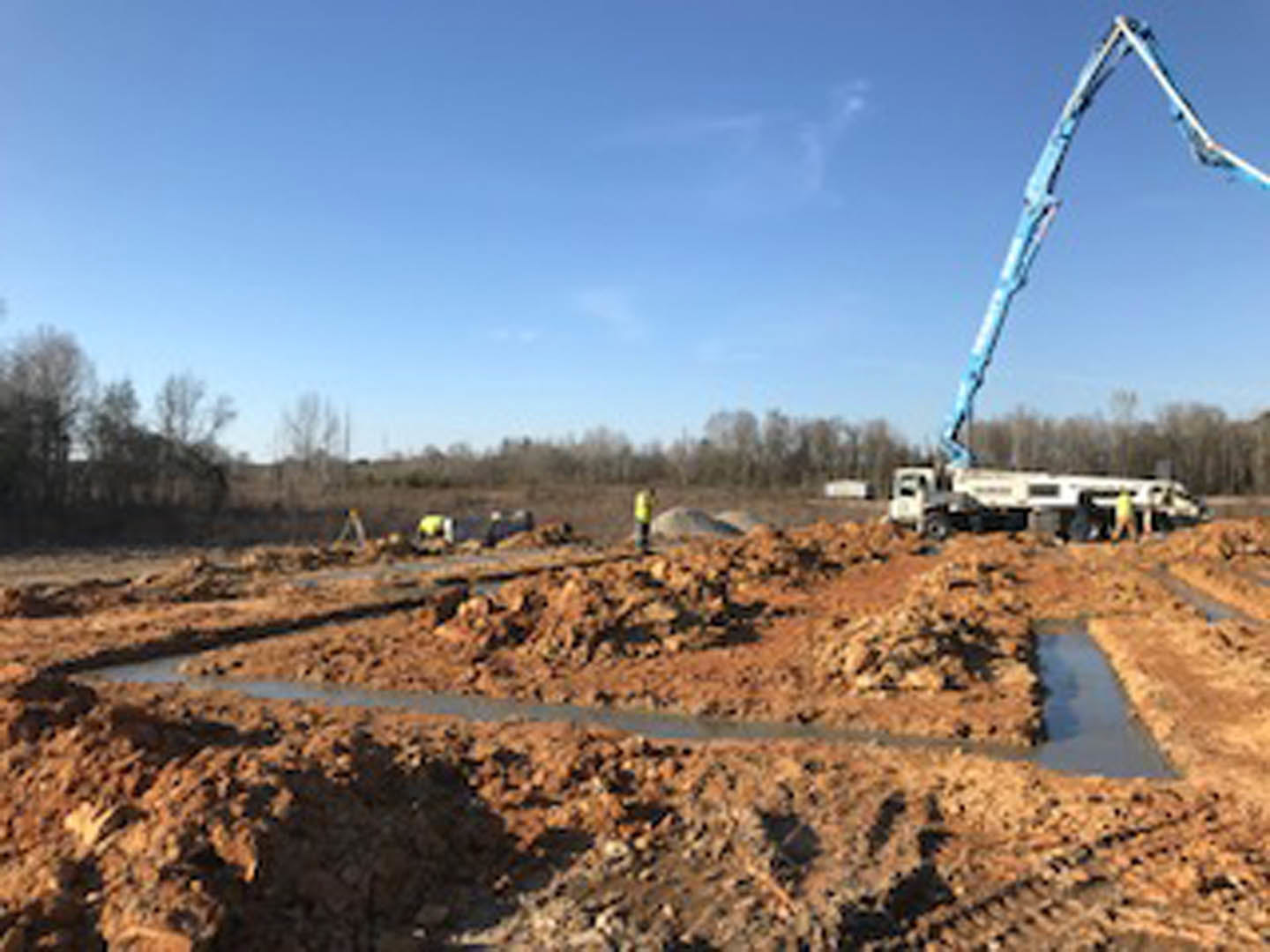Crane lifting a pole at custom home construction site with dirt road, white vehicle, blue sky, and scattered trees