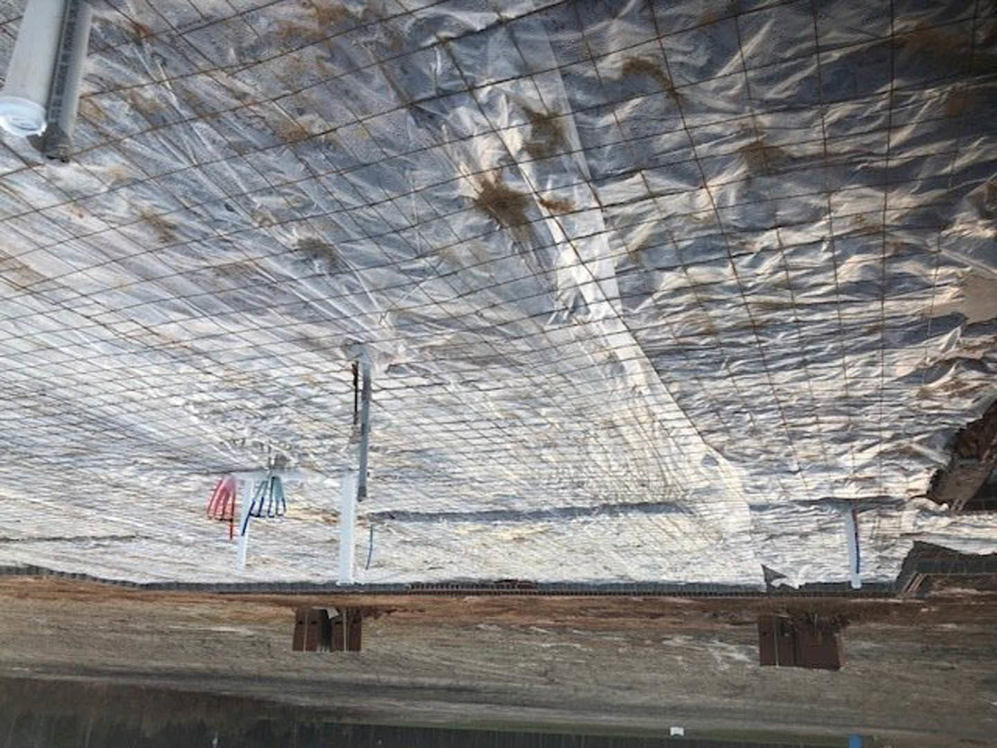 White tarp draped over wooden fence outdoors, partially obscuring view; metal tube visible in foreground, with blurred hand and fingers near edge of frame.