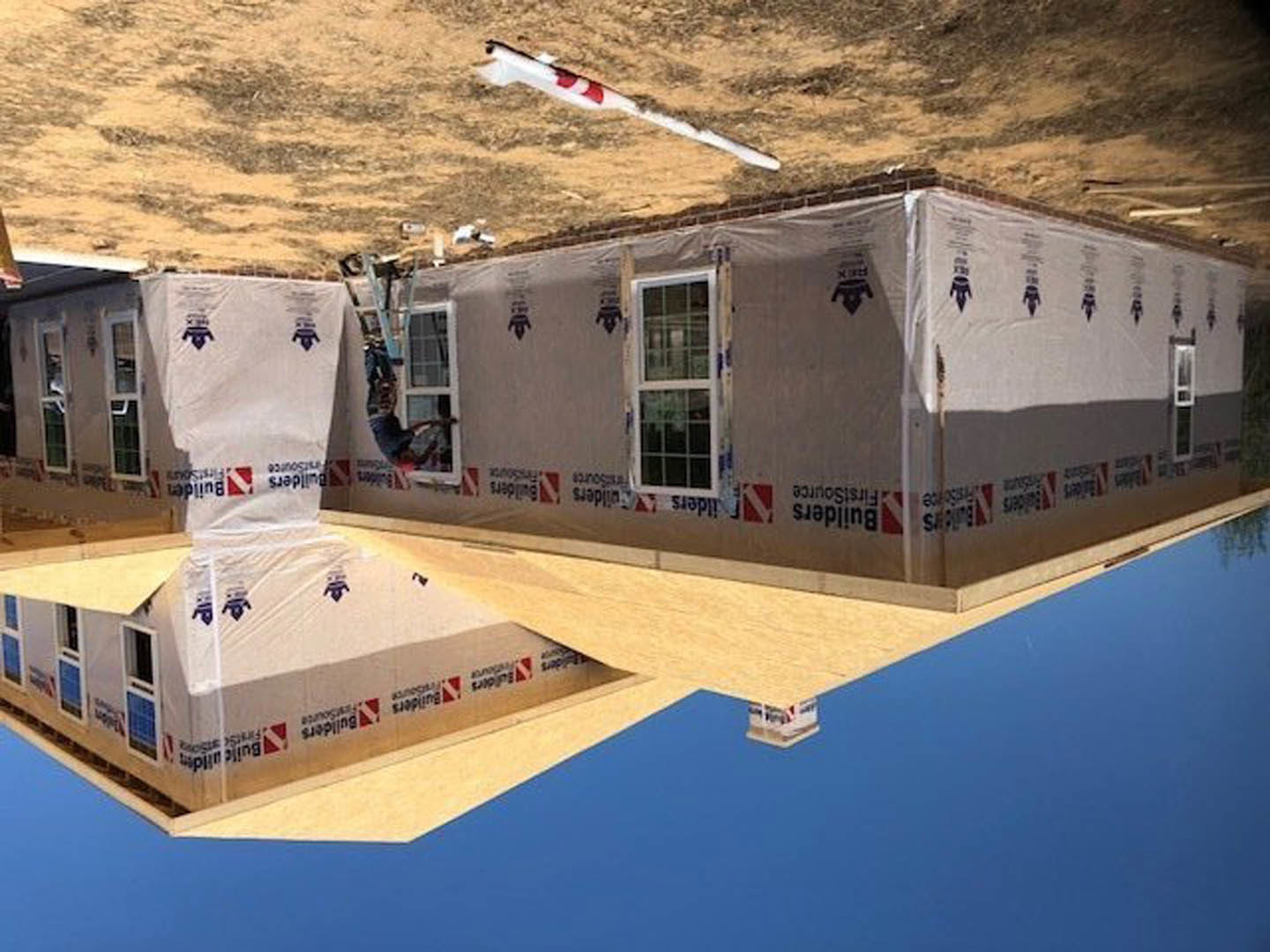 Modern home under construction with exposed framing, large crane lifting materials, and tall metal pole against clear sky