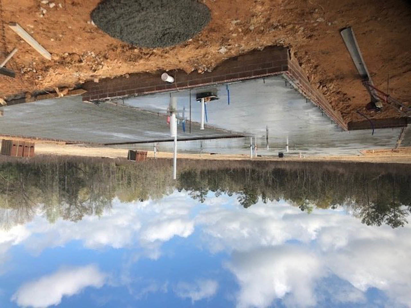 Concrete foundation topped with red brick wall, surrounded by brick base, outdoor setting with cloudy sky reflected in nearby water.
