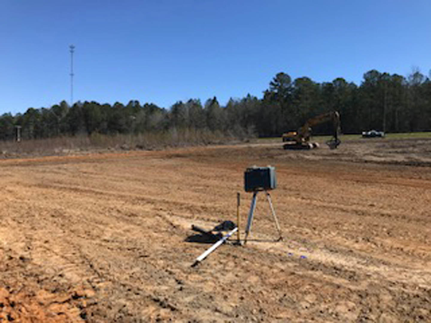 Camera on tripod set up in open dirt field with patches of grass, surrounded by trees under clear blue sky