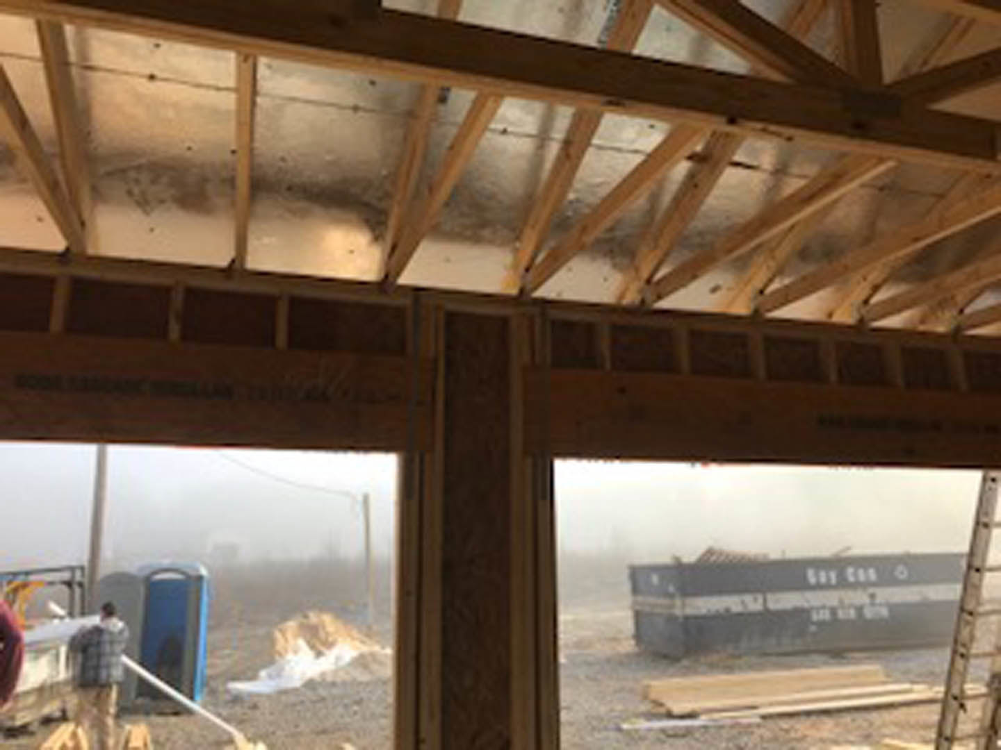 Open-concept interior with exposed wooden ceiling beams, blurred figure walking near large windows, partial view of exterior building and sky through glass