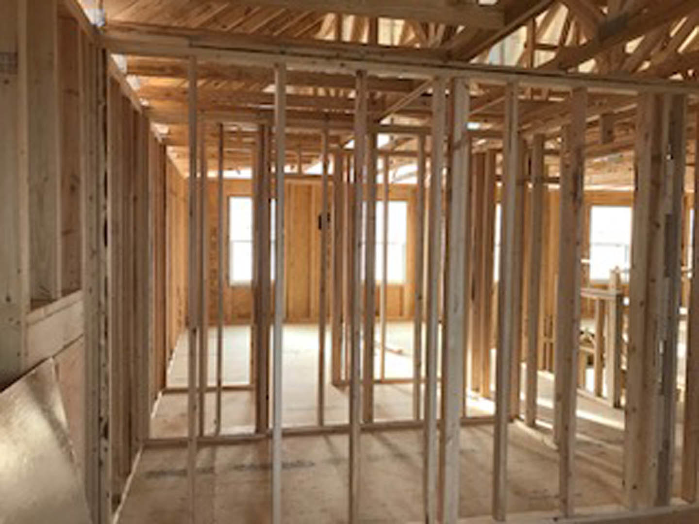 Wood-framed room under construction with exposed beams, unfinished floor, and daylight streaming through a window; blurred hand visible in foreground.