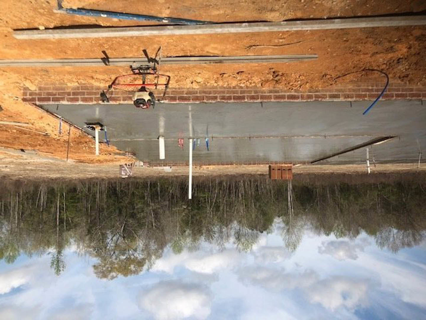 Framed custom home under construction with exposed wooden framing, dirt ground, and construction equipment in foreground
