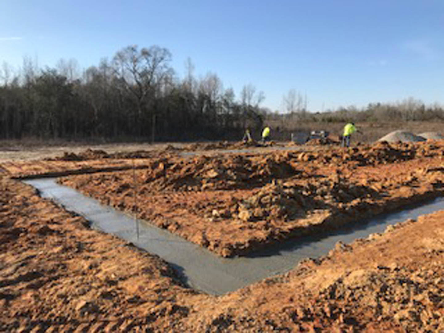 Men working on dirt foundation at residential construction site, surrounded by soil, mud, and scattered building materials; blue sky and trees in background.