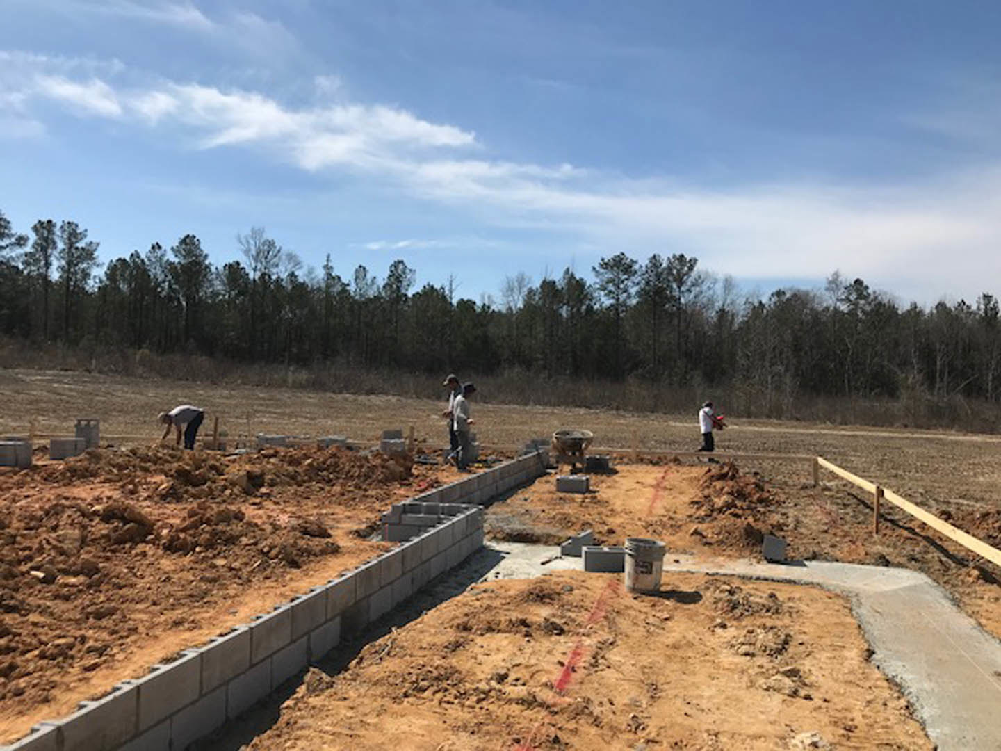 Workers assembling foundation forms on a grassy construction site with exposed soil, surrounded by trees and a cloudy sky