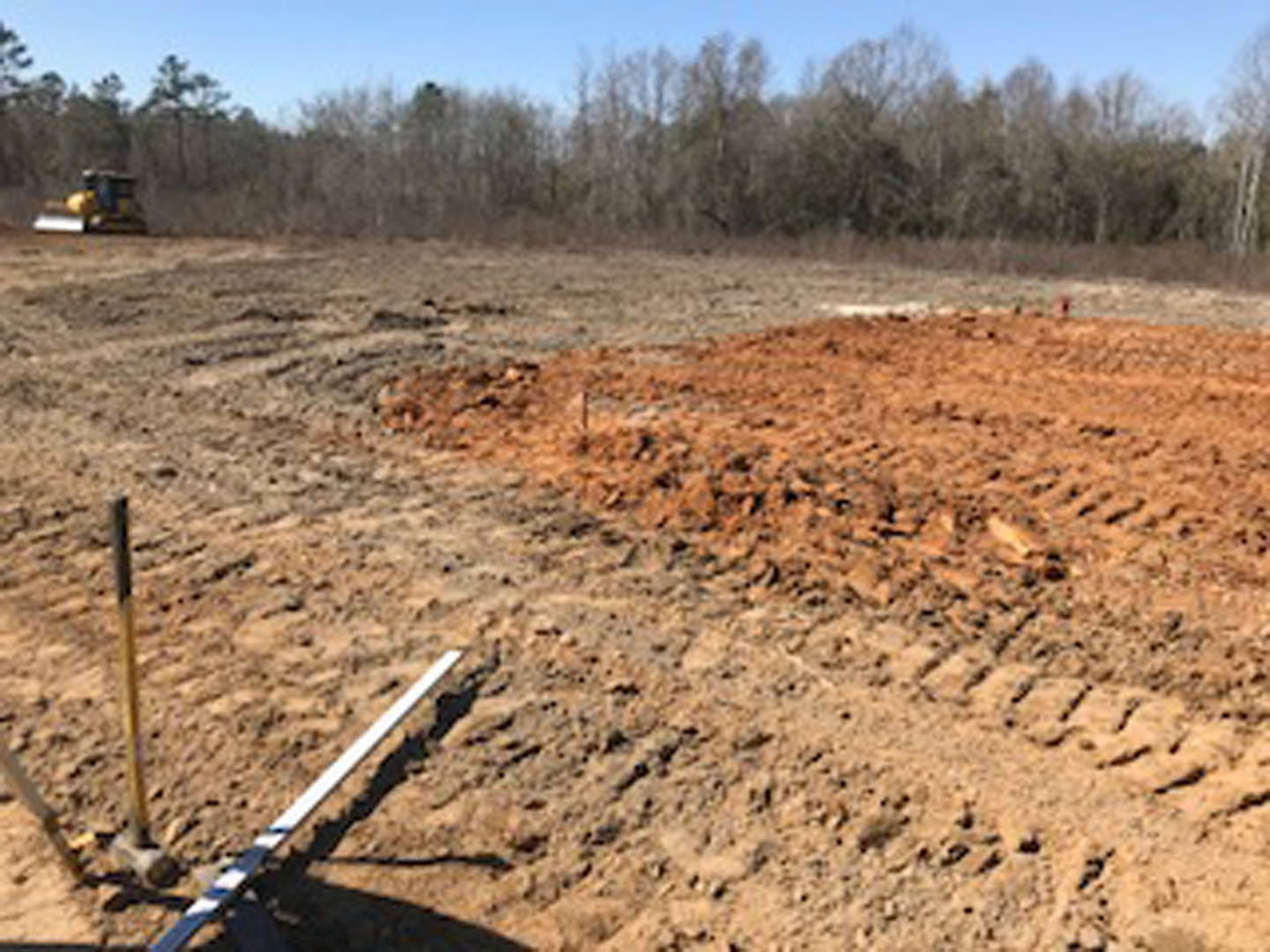 Dirt field with two mounds of soil, metal pole, scattered vegetation, and trees in the background under a clear sky