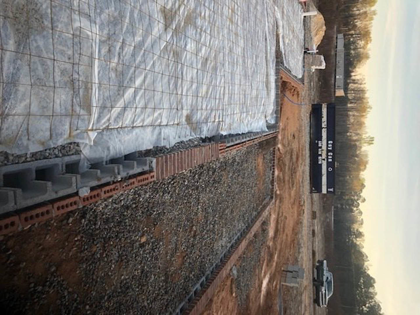 Gravel-covered construction site with a parked truck, white mesh draped over a partially built wall, distant group of trees, and scattered signage