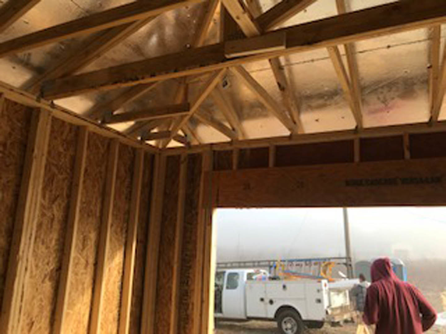 Exposed wooden framing of a house under construction, person in red hoodie near white van with ladder, group of people standing by white truck, close-up of tire and blurry leg in