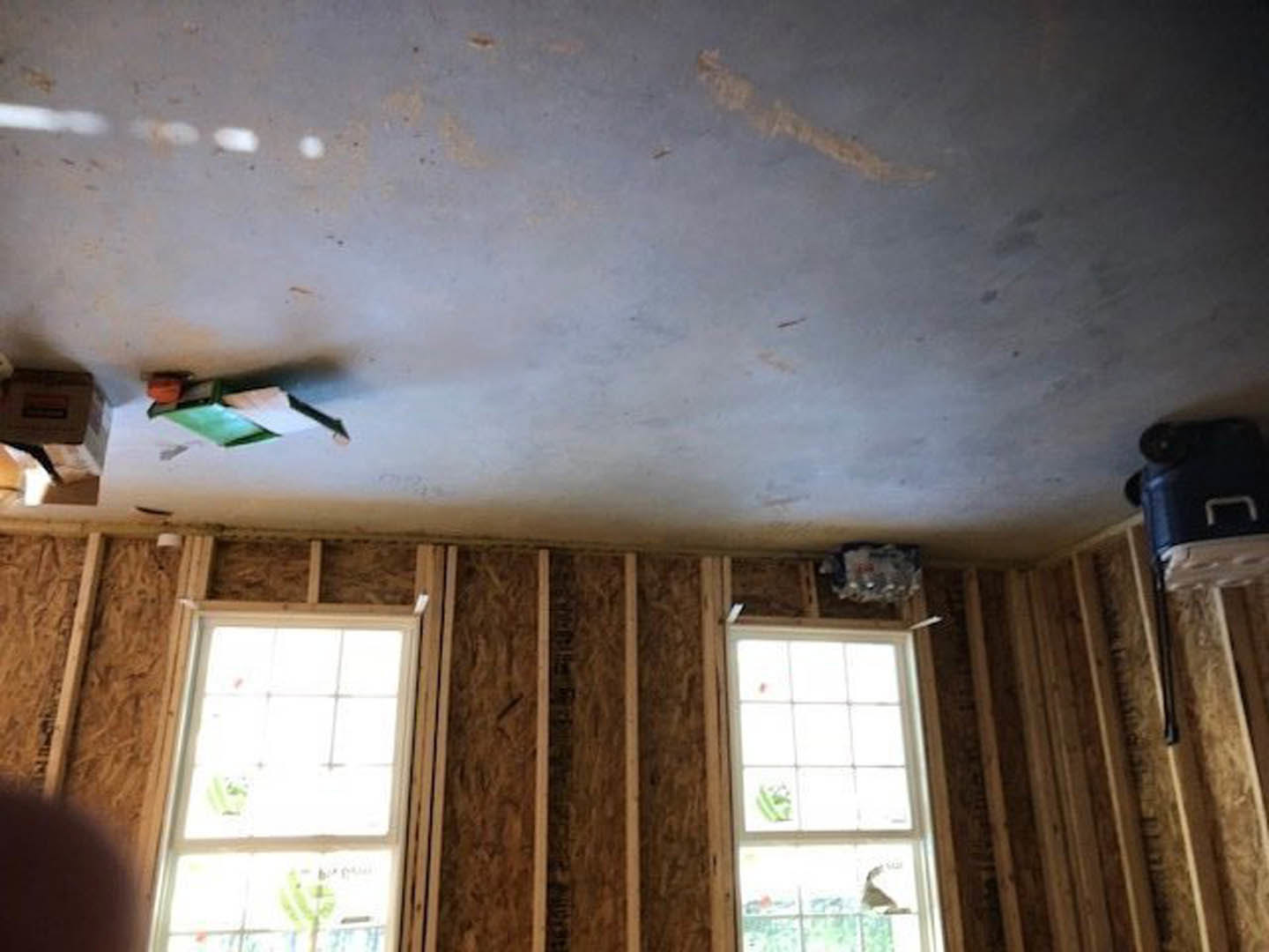 Bright room with white plaster ceiling, large window featuring multiple square panes and white frame, wood-framed wall, sheer curtains, and blue sky visible outside.