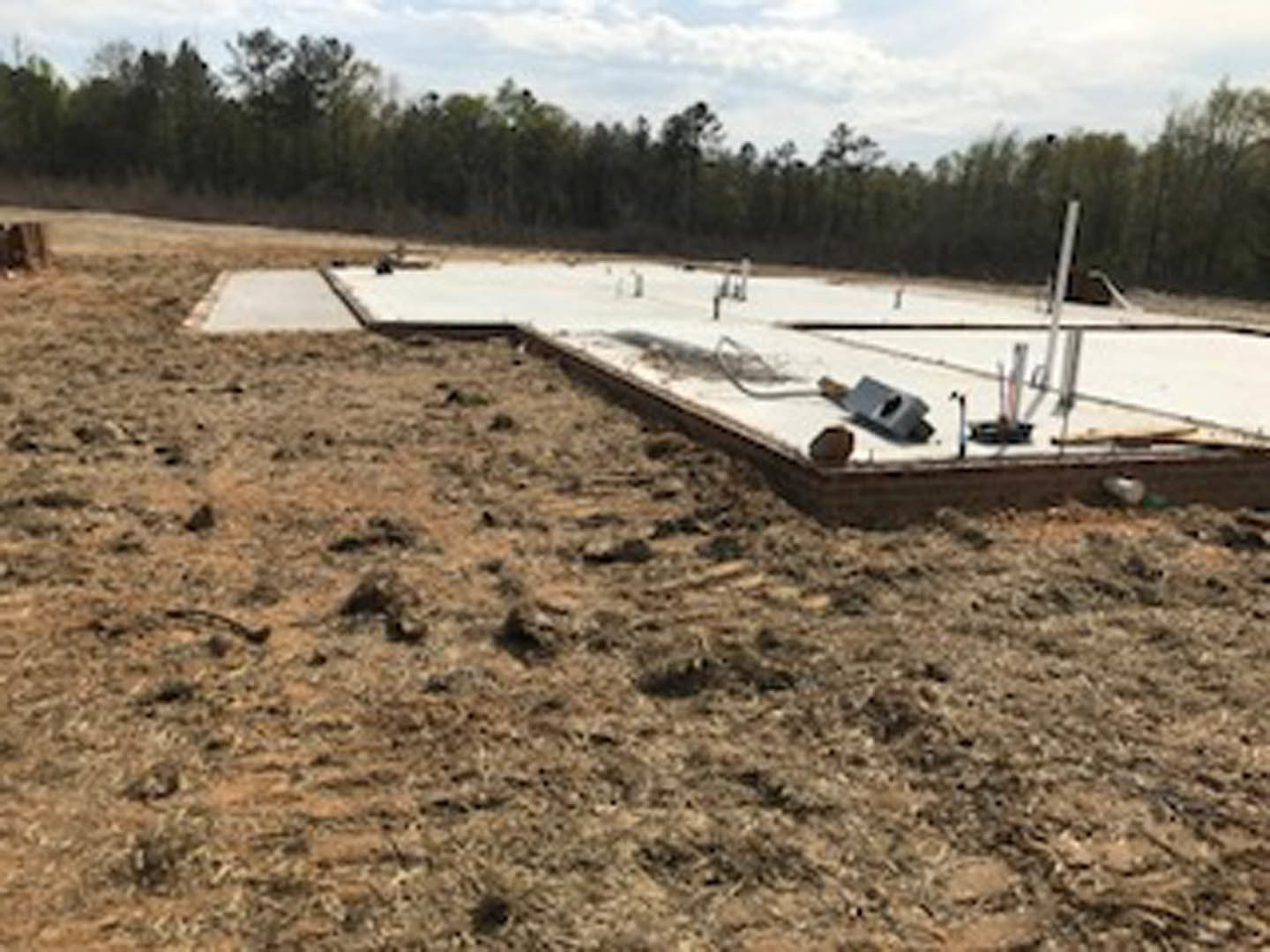 Concrete foundation slab with several metal poles set in an open grassy field, surrounded by trees and cloudy sky.